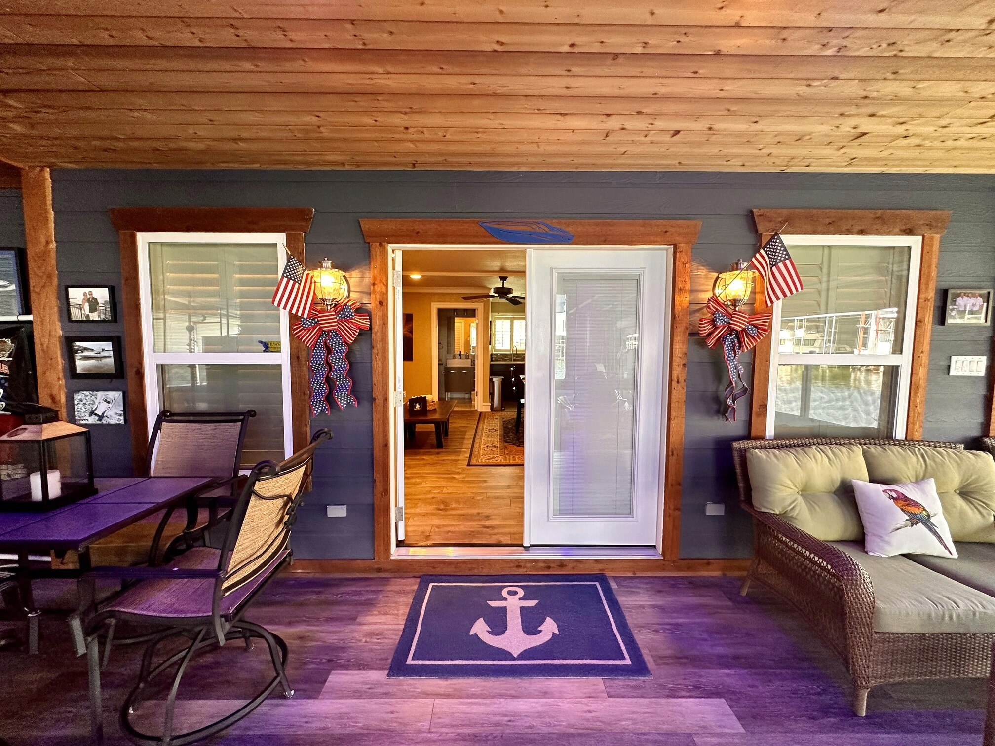 A cozy, covered porch with rustic wooden ceiling and navy blue walls features a set of wicker and metal furniture, decorated with American flags and nautical elements, such as anchor symbols on rugs, exuding a charming, patriotic atmosphere.
