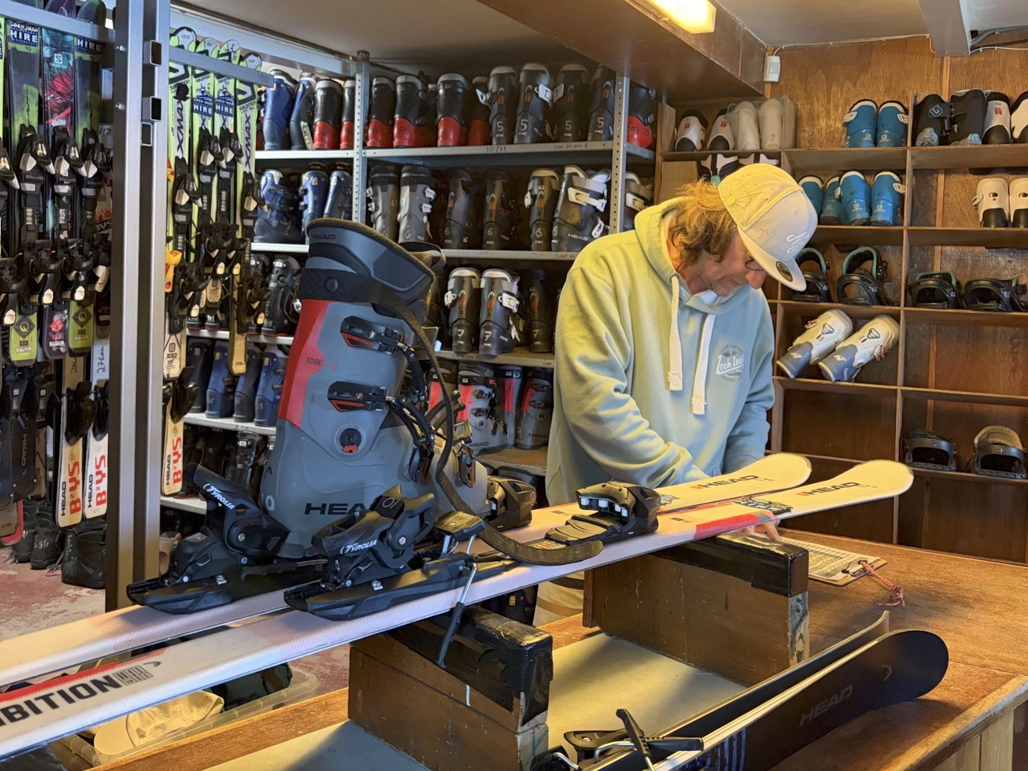 A person in a hoodie and cap adjusts a ski on a workbench in a ski rental shop. Shelves with ski boots and equipment surround them, creating a focused atmosphere.