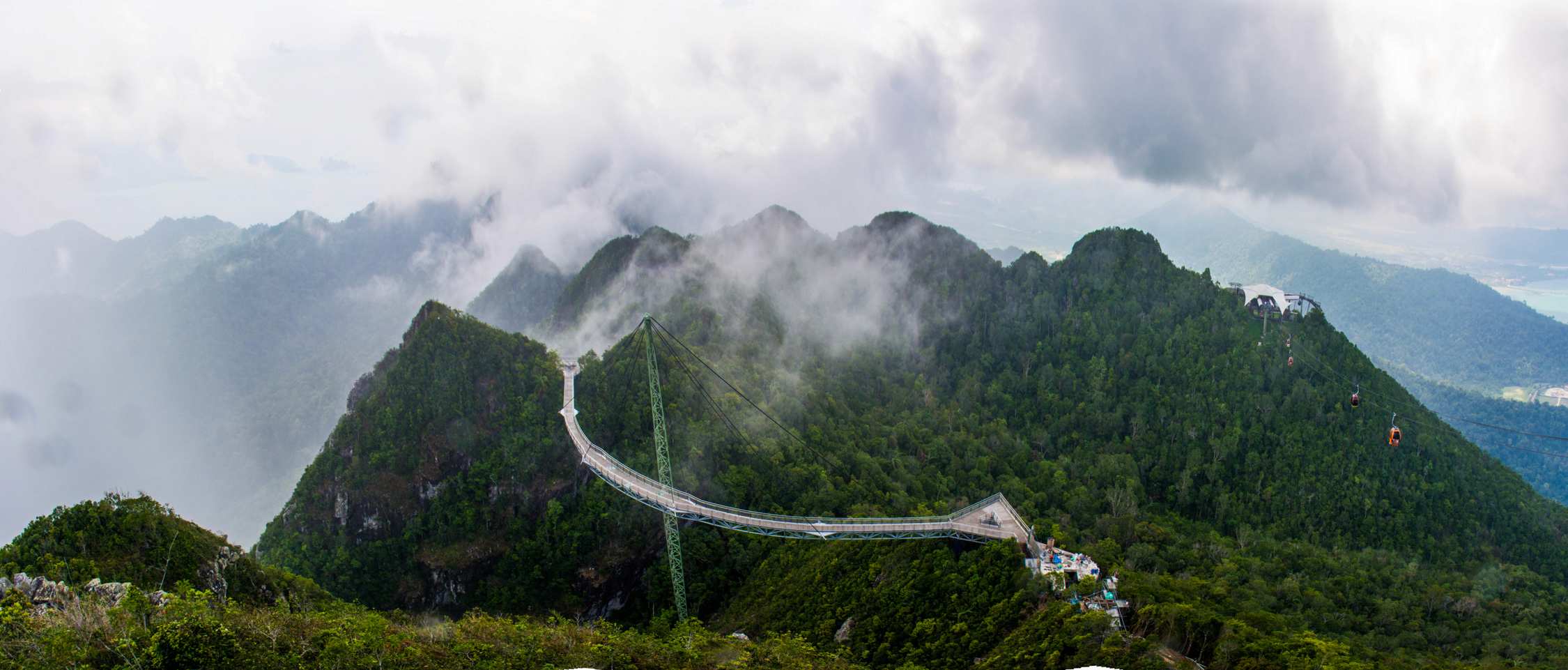 A winding mountain road curves through lush green peaks under a cloudy sky with mist clinging to the slopes.