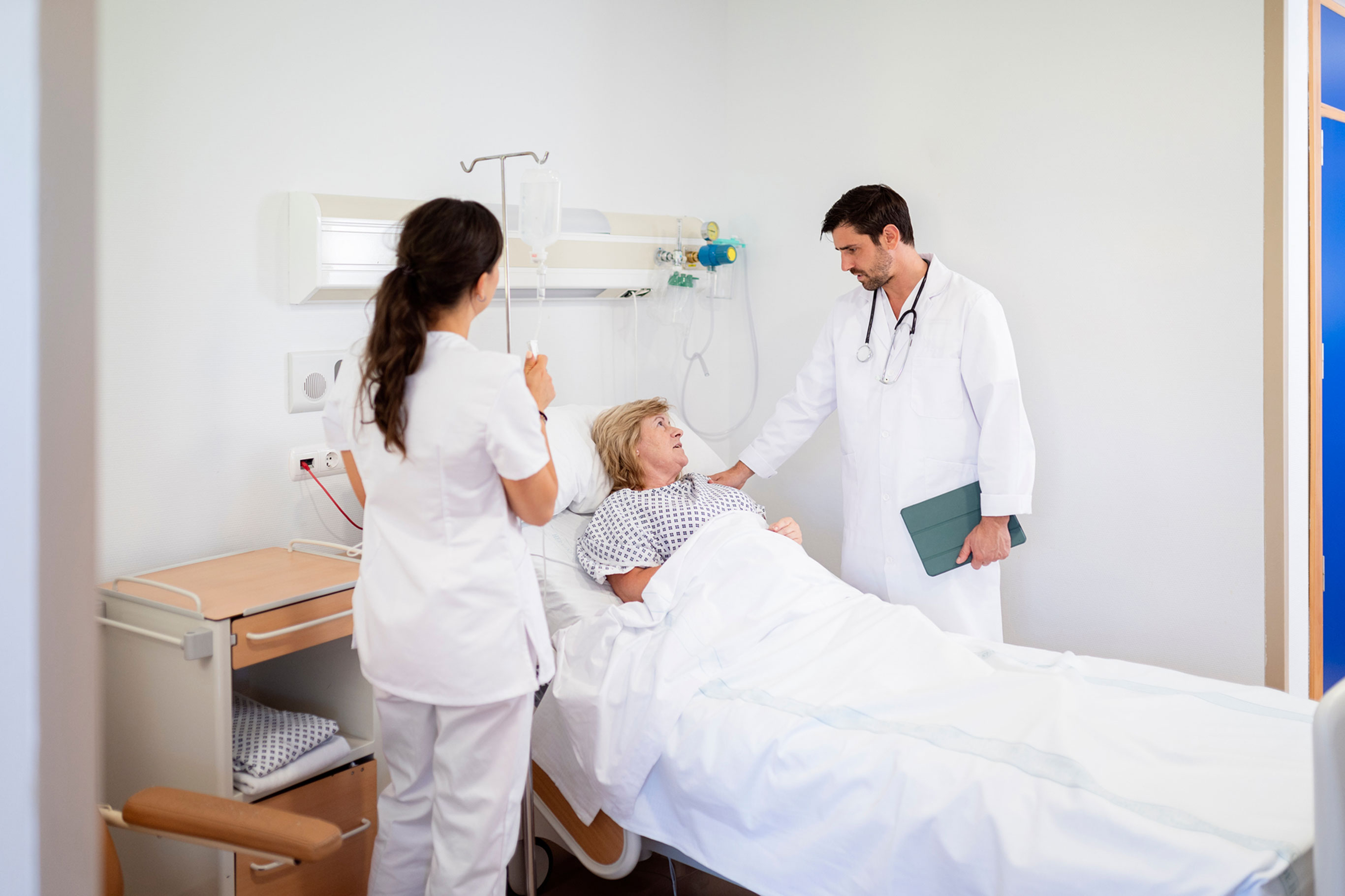 A doctor in a white lab coat and a nurse in white scrubs attend to a patient lying in a hospital bed.