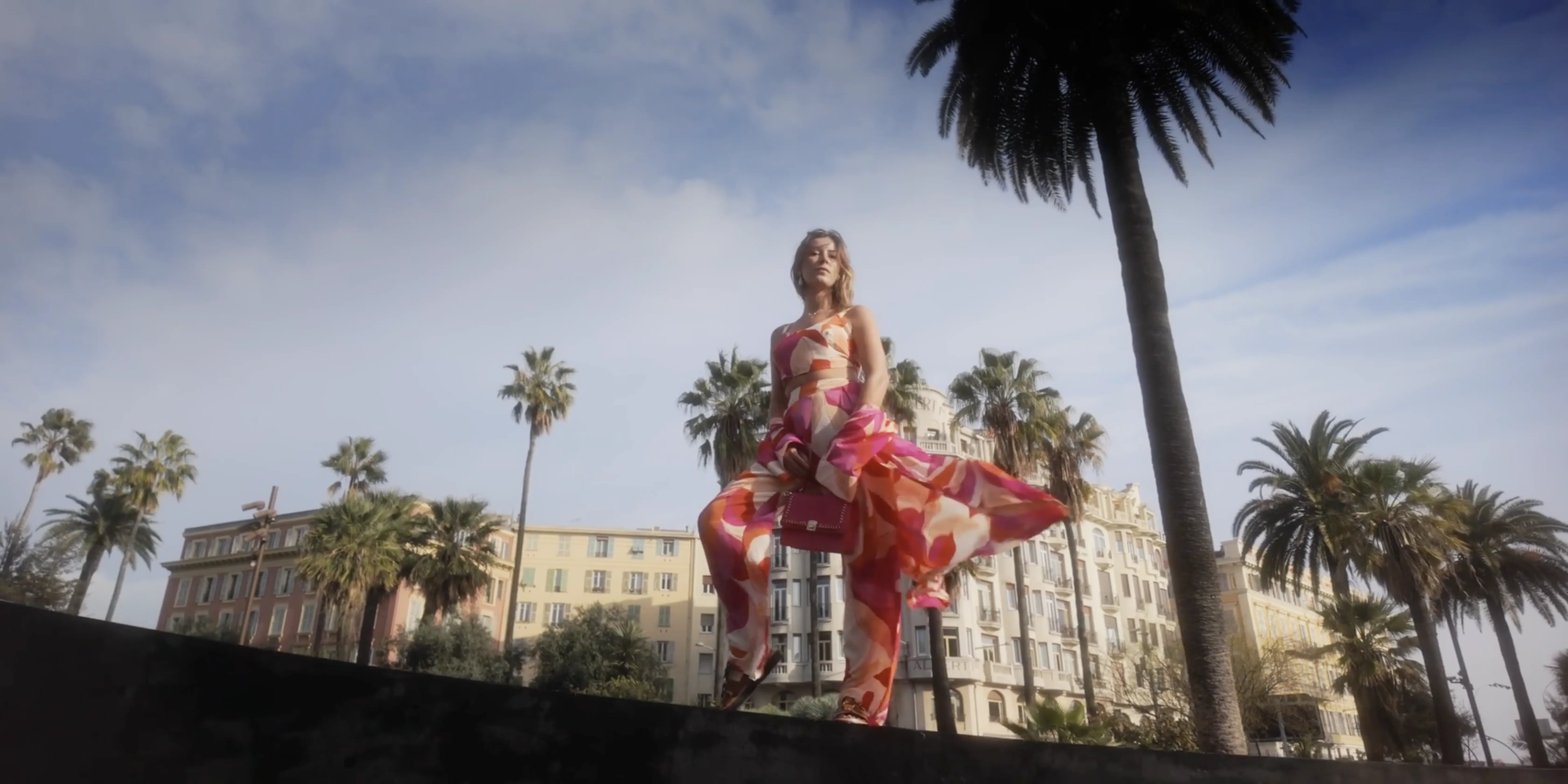 A woman in a flowing, vibrant pink and orange dress joyfully dances on a ledge, surrounded by tall palm trees against a backdrop of historic buildings and a partially cloudy blue sky.