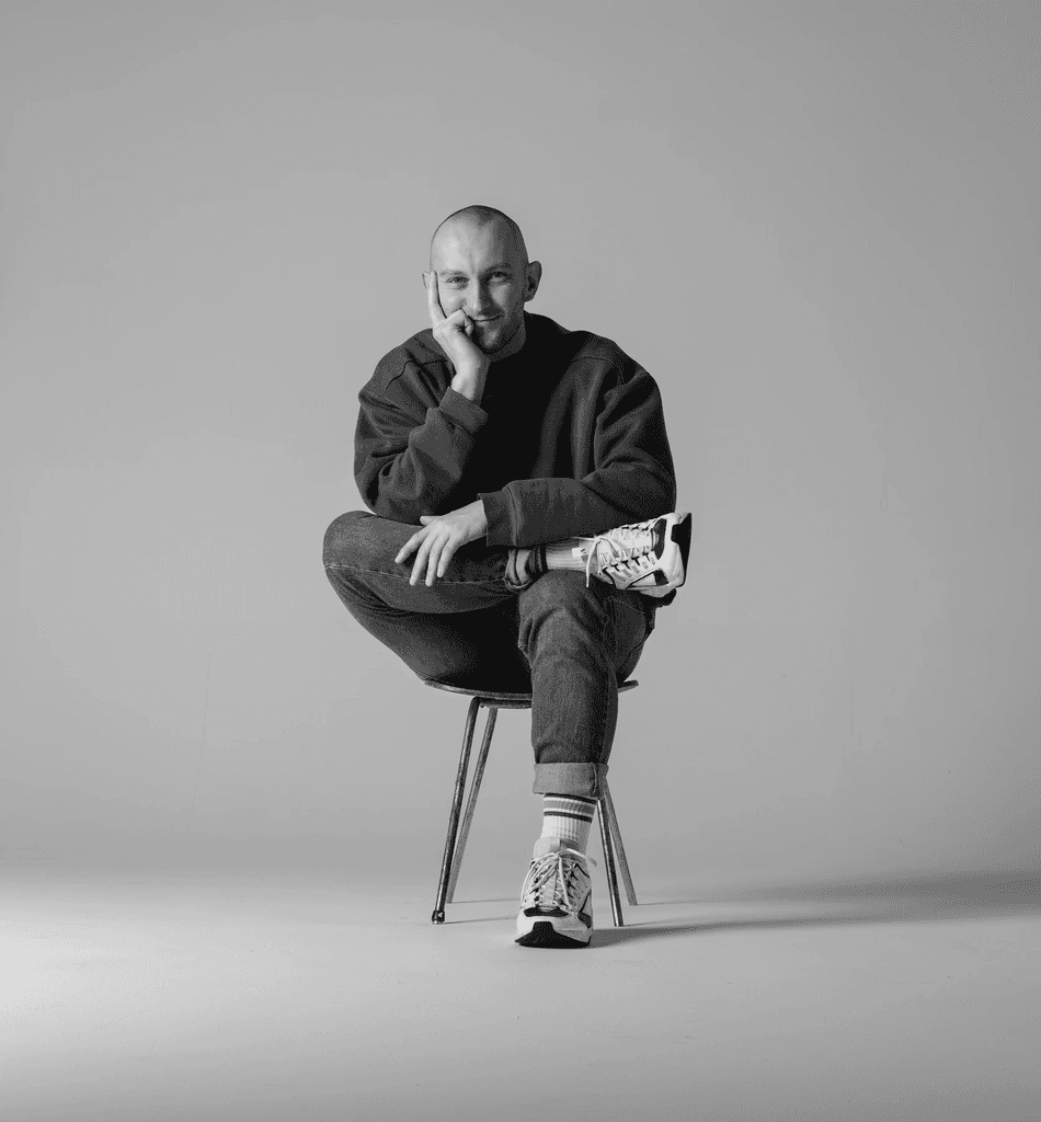 Close-up portrait of a person wearing a gray insulated hooded jacket with the hood up, showing their face framed by the hood against a light background.