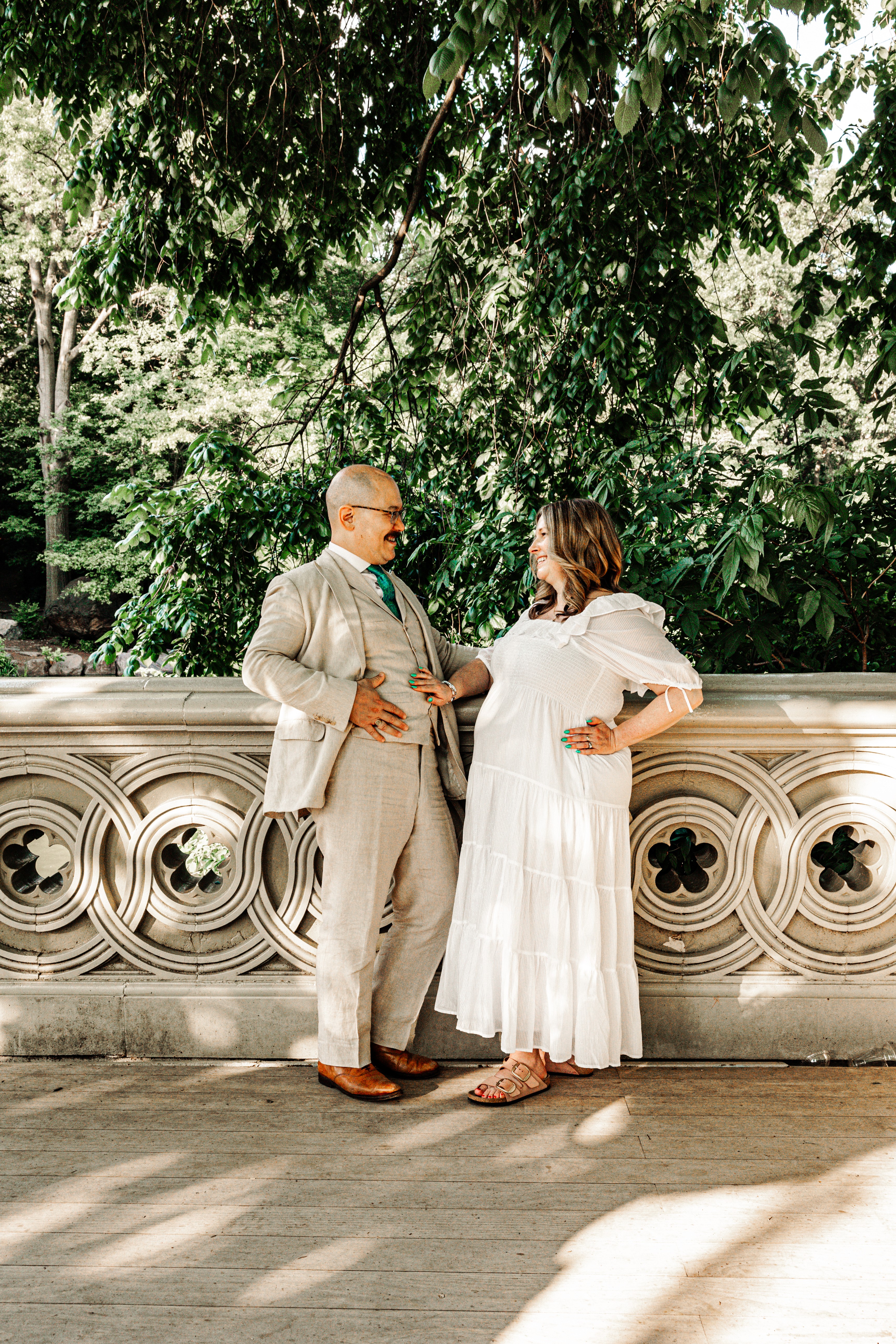 Expecting couple sharing a joyful, laughing moment on Bow Bridge in Central Park, NYC — he's dapper in a linen suit, she's glowing in a white dress, both cradling her baby bump together — natural, candid maternity photography by Lizz Spano Photography, New York City maternity photographer.