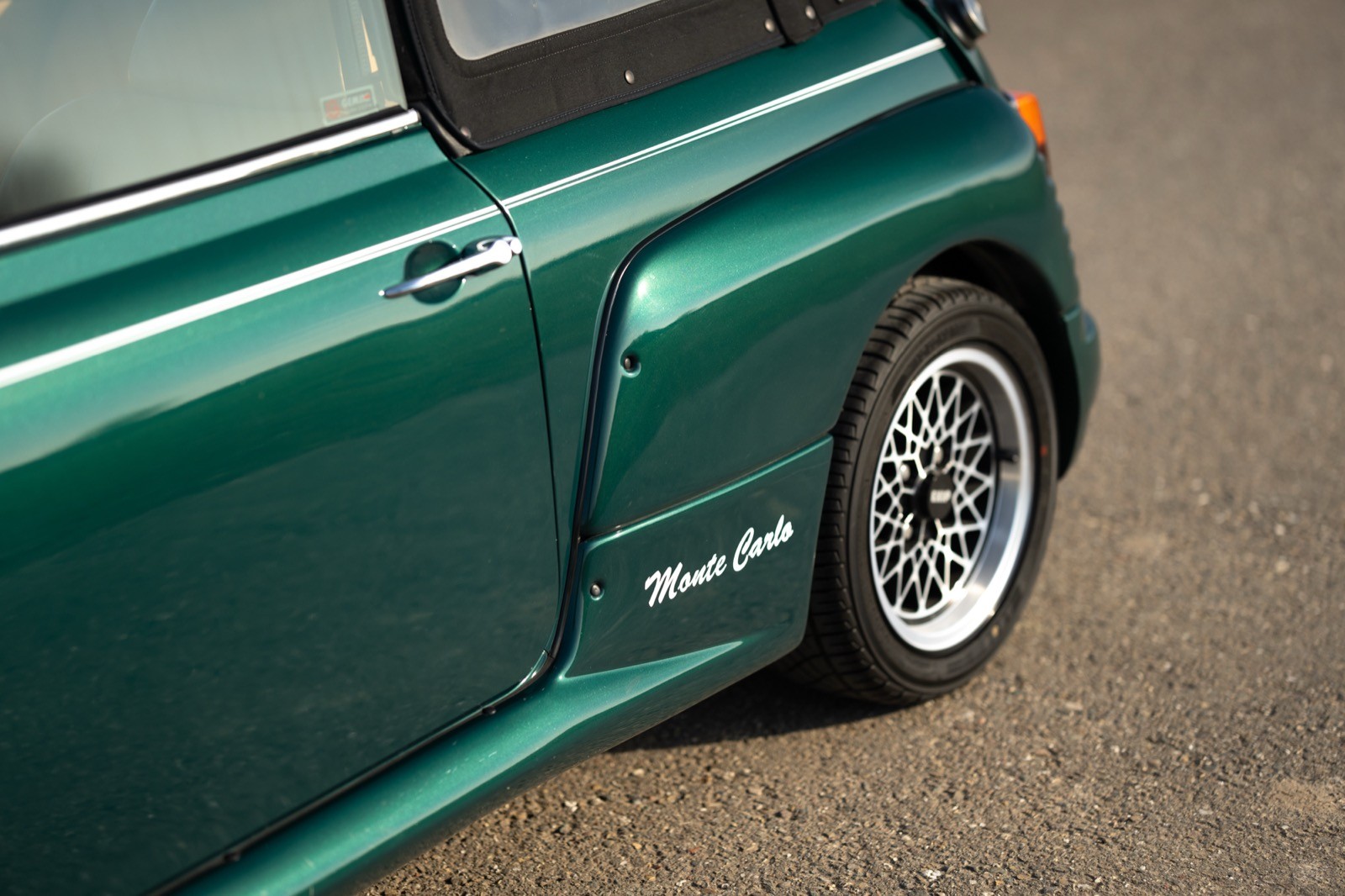 Close-up of a green car's side, showcasing its wheel, fender, and sleek body design on a textured surface.