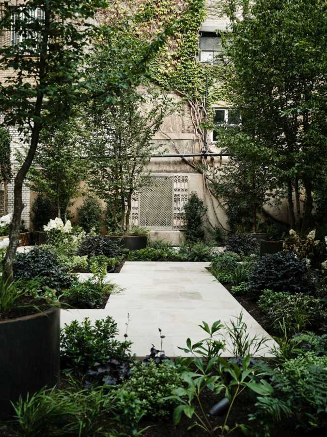 Architectural courtyard landscaping in London, showcasing a minimalist light stone pathway winding through lush green shrubbery and mature trees adjacent to a residential building.