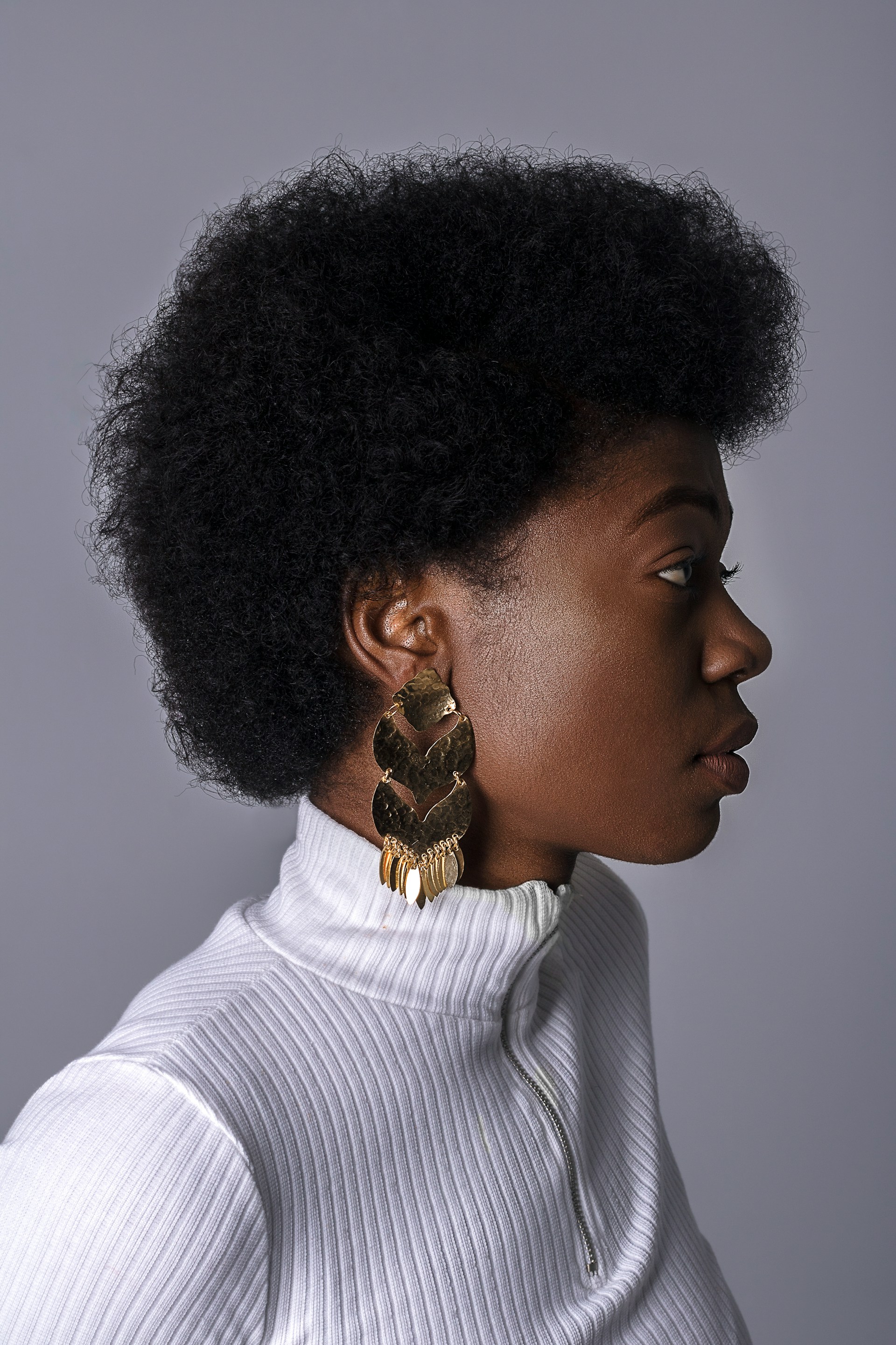Side profile of a young Black woman with a natural, rounded afro, wearing a white ribbed turtleneck and a large, multi-tiered gold earring against a grey studio backdrop.