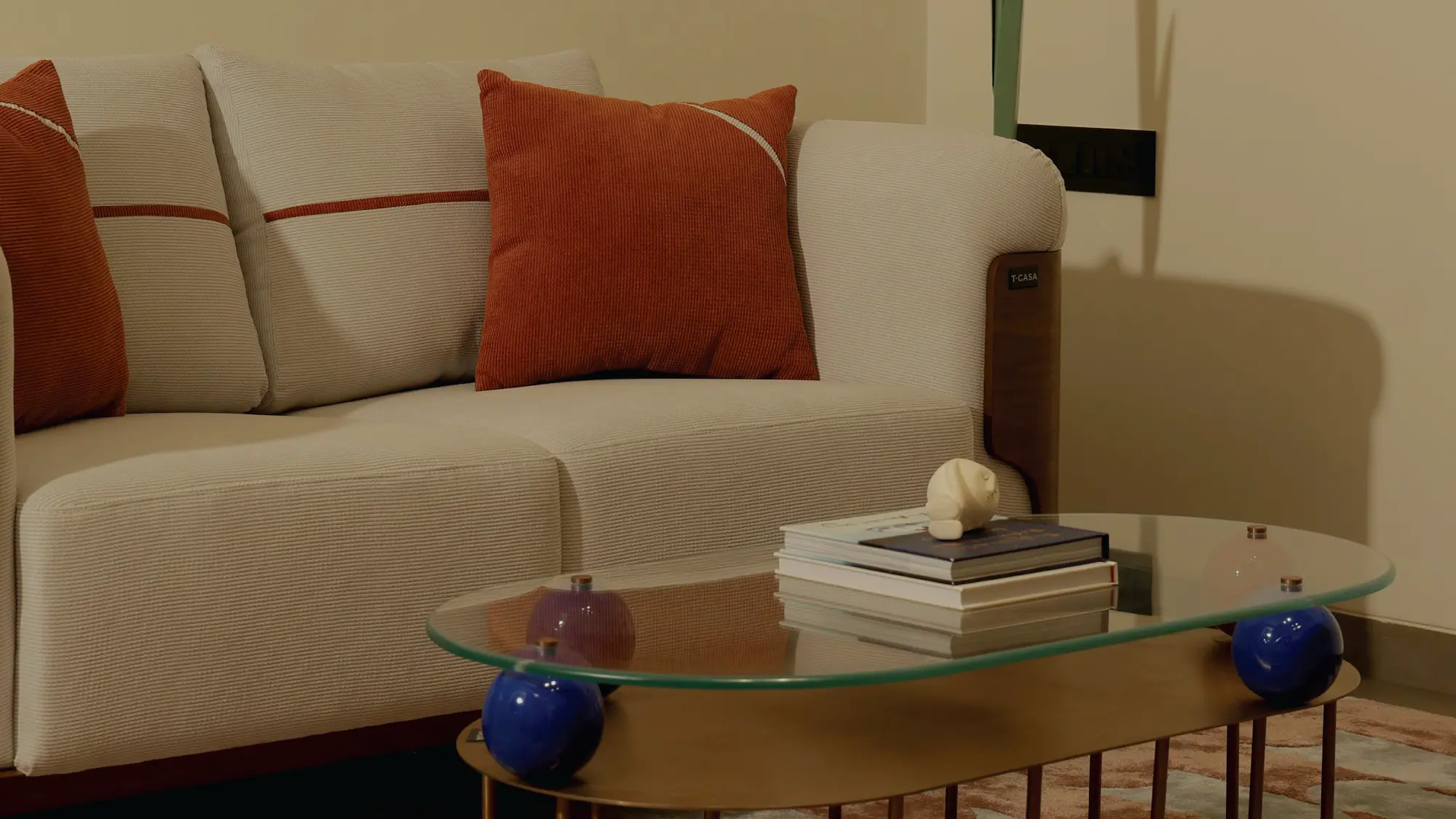 A modern living room features a light beige sofa with two plush, burnt orange cushions and a stylish glass coffee table adorned with a small stack of books and a decorative ornament.