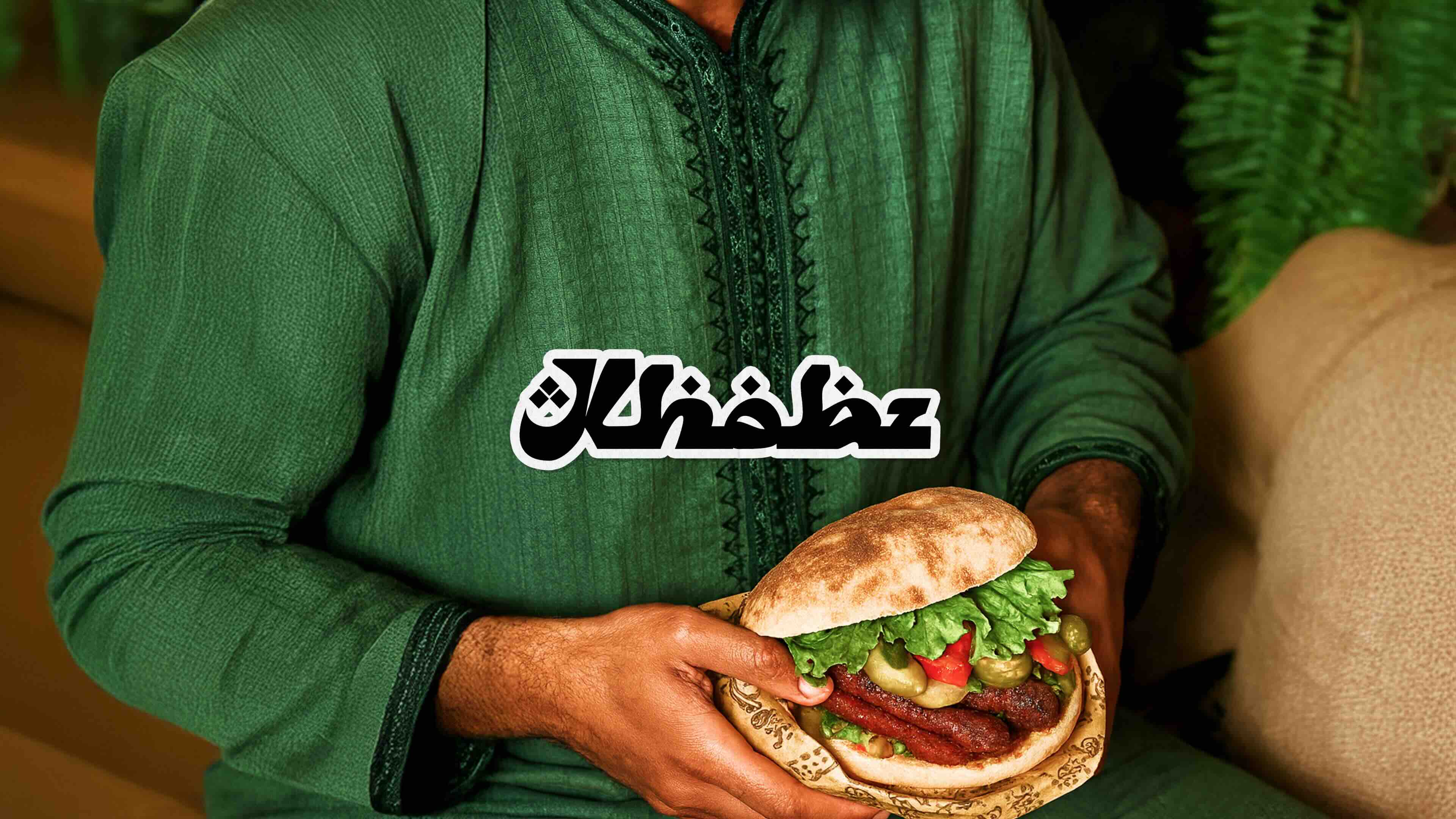 Smiling person holding a large burger with fried chicken, cheese, lettuce, tomato, pickles, and sauce, about to take a bite.