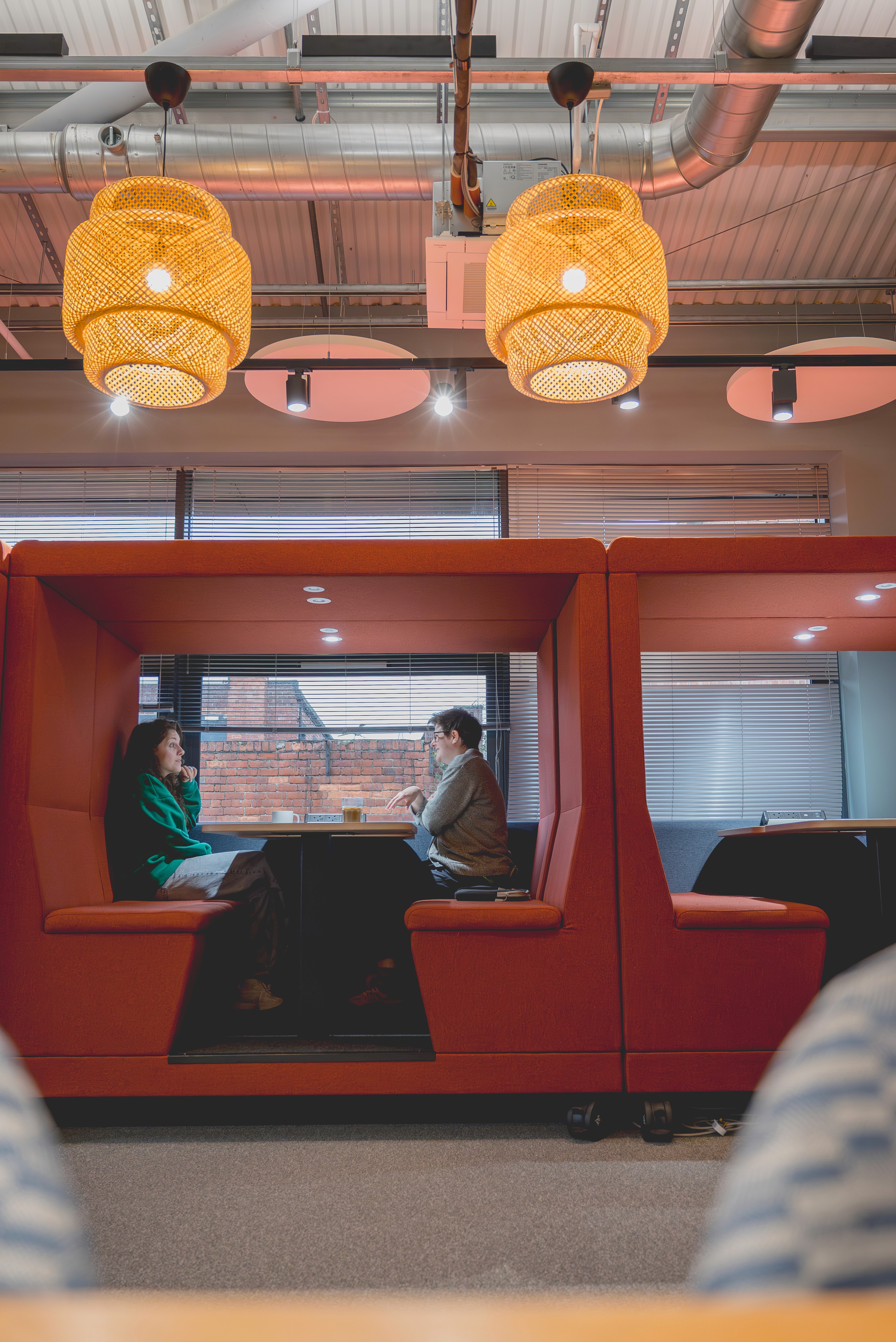 Two people sit across from each other in a booth-style seating area within an office lounge, with warm pendant lights hanging overhead and large windows behind them.