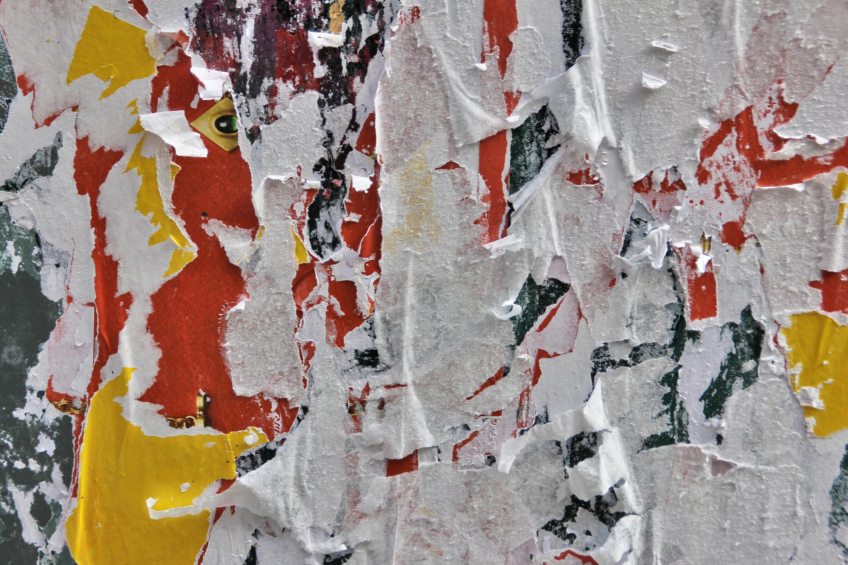 Close-up photograph of peeling posters revealing layers of red, yellow, white, and black paper. A faint eye peers through the torn surface, creating a haunting interplay between concealment, exposure, and time-worn texture.