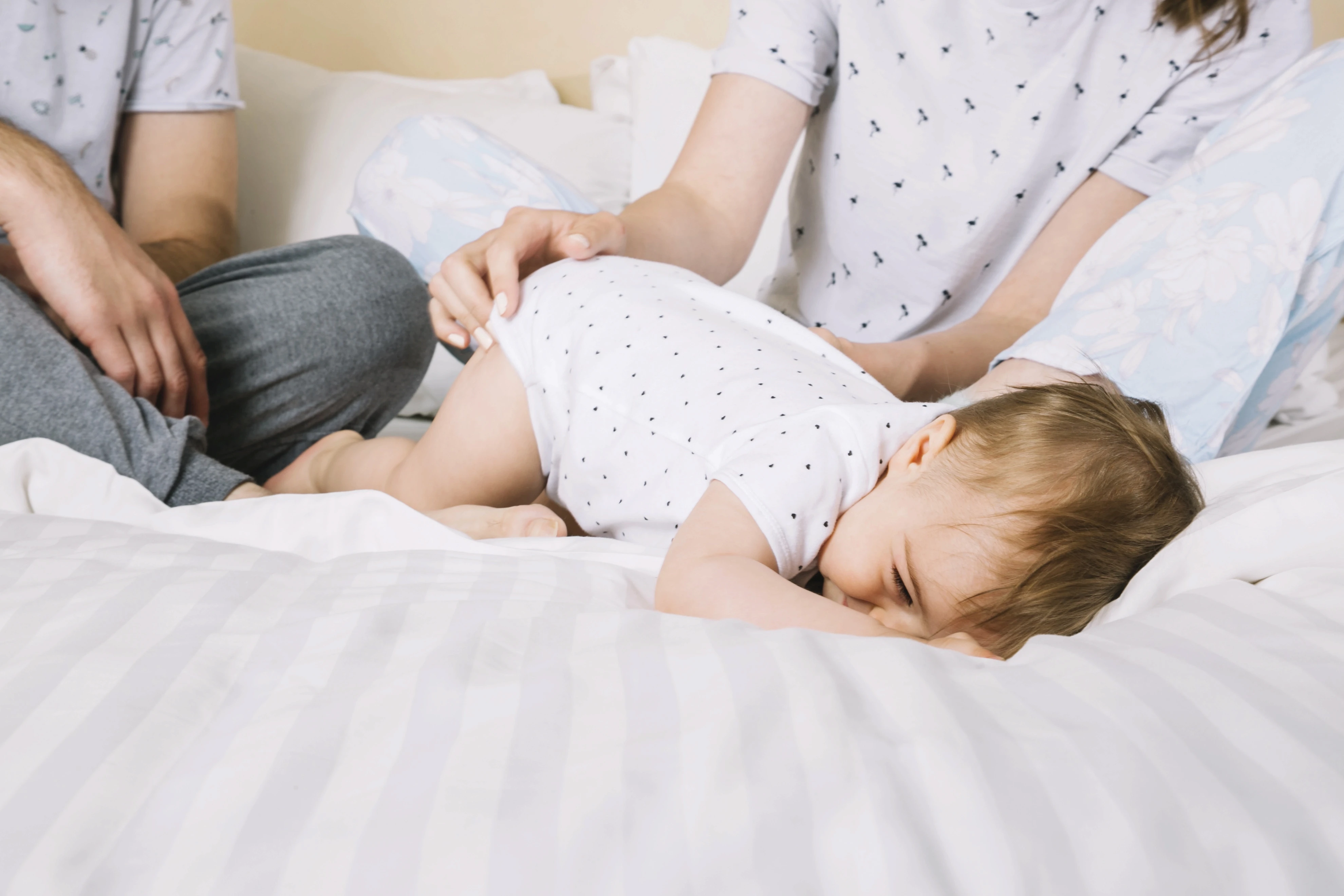 Um bebê pequeno deitado de bruços em uma cama com lençóis brancos, com o rosto virado para o lado e chorando ou incomodado, sugerindo dor ou cólica. As mãos de dois adultos (os pais) são visíveis na cena, observando o bebê.
