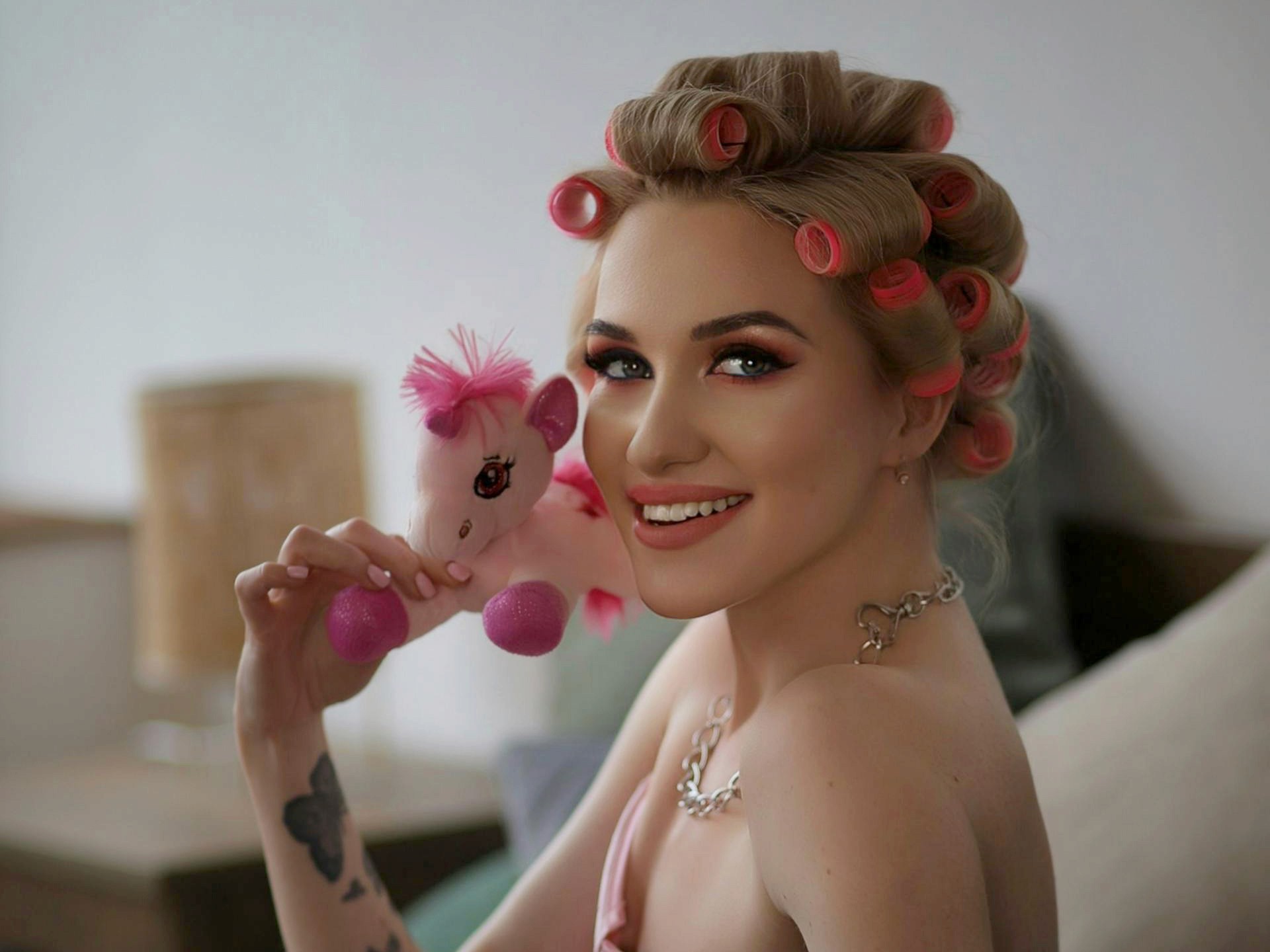 A smiling blonde woman with pink hair rollers and pink makeup, holding a small pink stuffed unicorn toy.