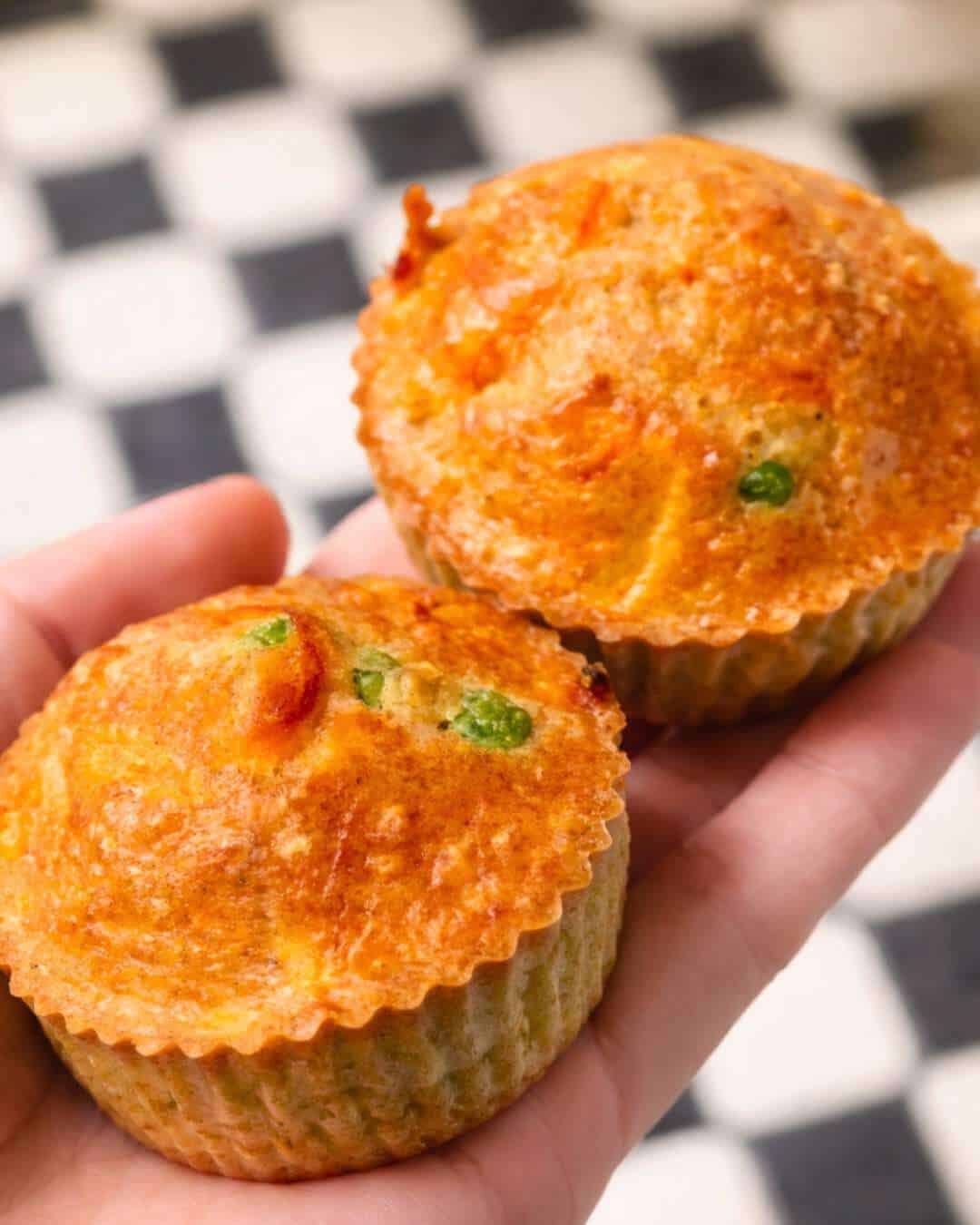 Two savory cheesy muffins held in a hand, shown close-up with a golden top and visible green vegetable pieces.