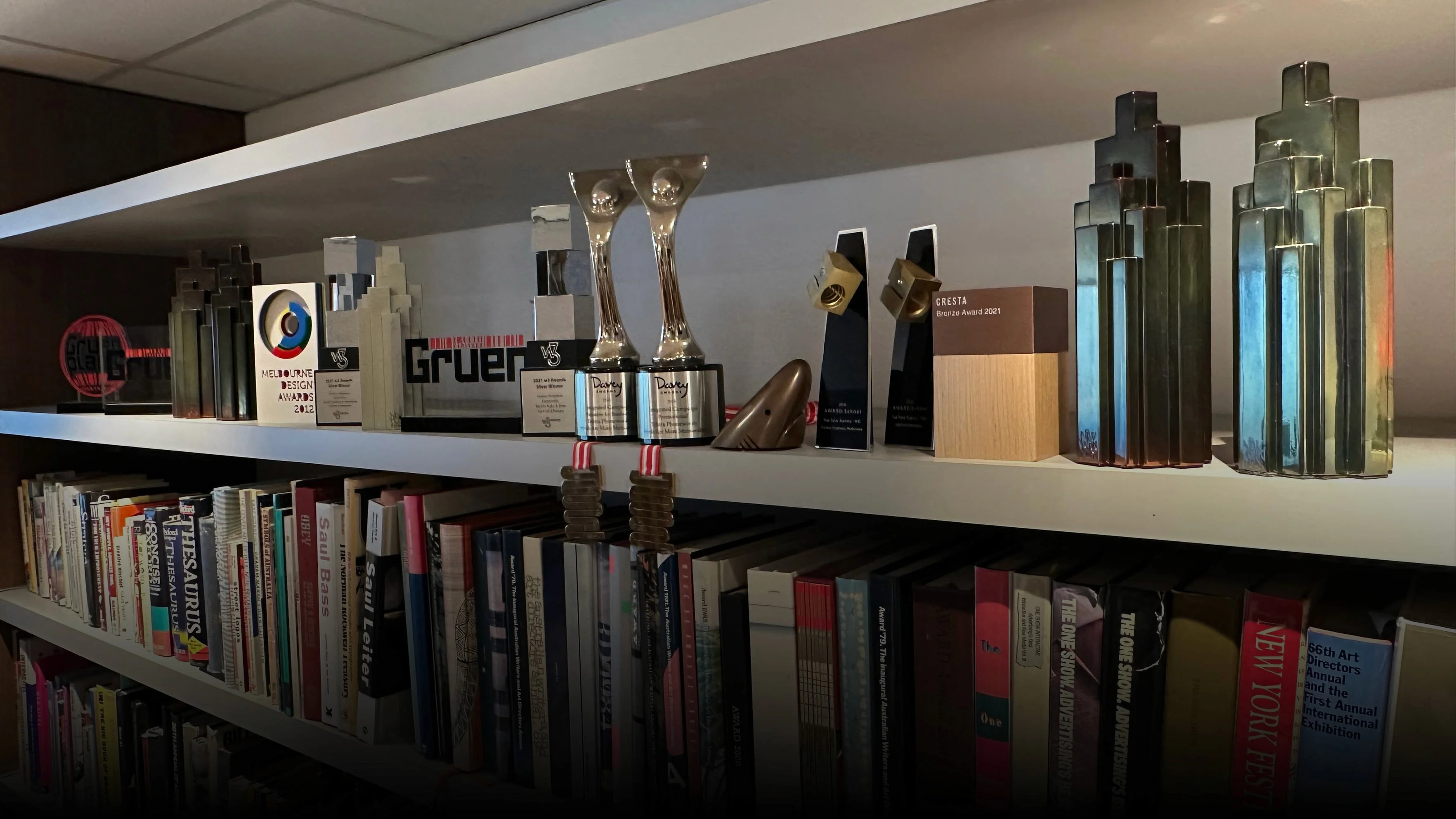 Shelf of agency award trophies in an office, including Davey, Gruen, Cresta and Melbourne Design Awards, above a row of books