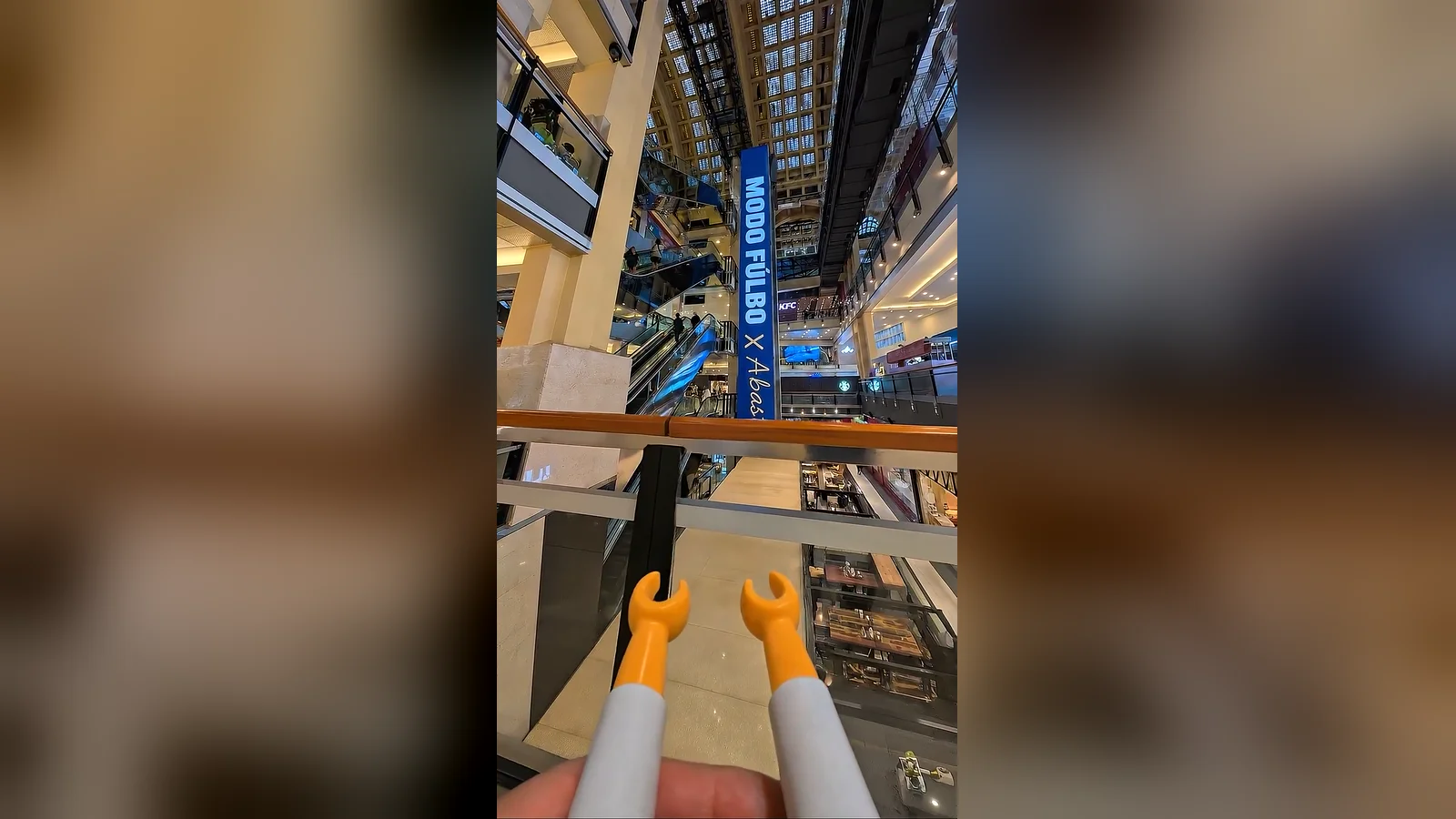 Manos amarillas de un Playmobil en primer plano frente al atrio y las escaleras mecánicas de Abasto Shopping