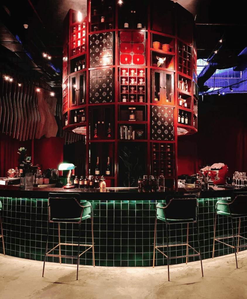 A modern bar interior featuring a circular structure with red cabinetry and shelves displaying bottles, accompanied by a sleek green-tiled bar counter and stylish bar stools.