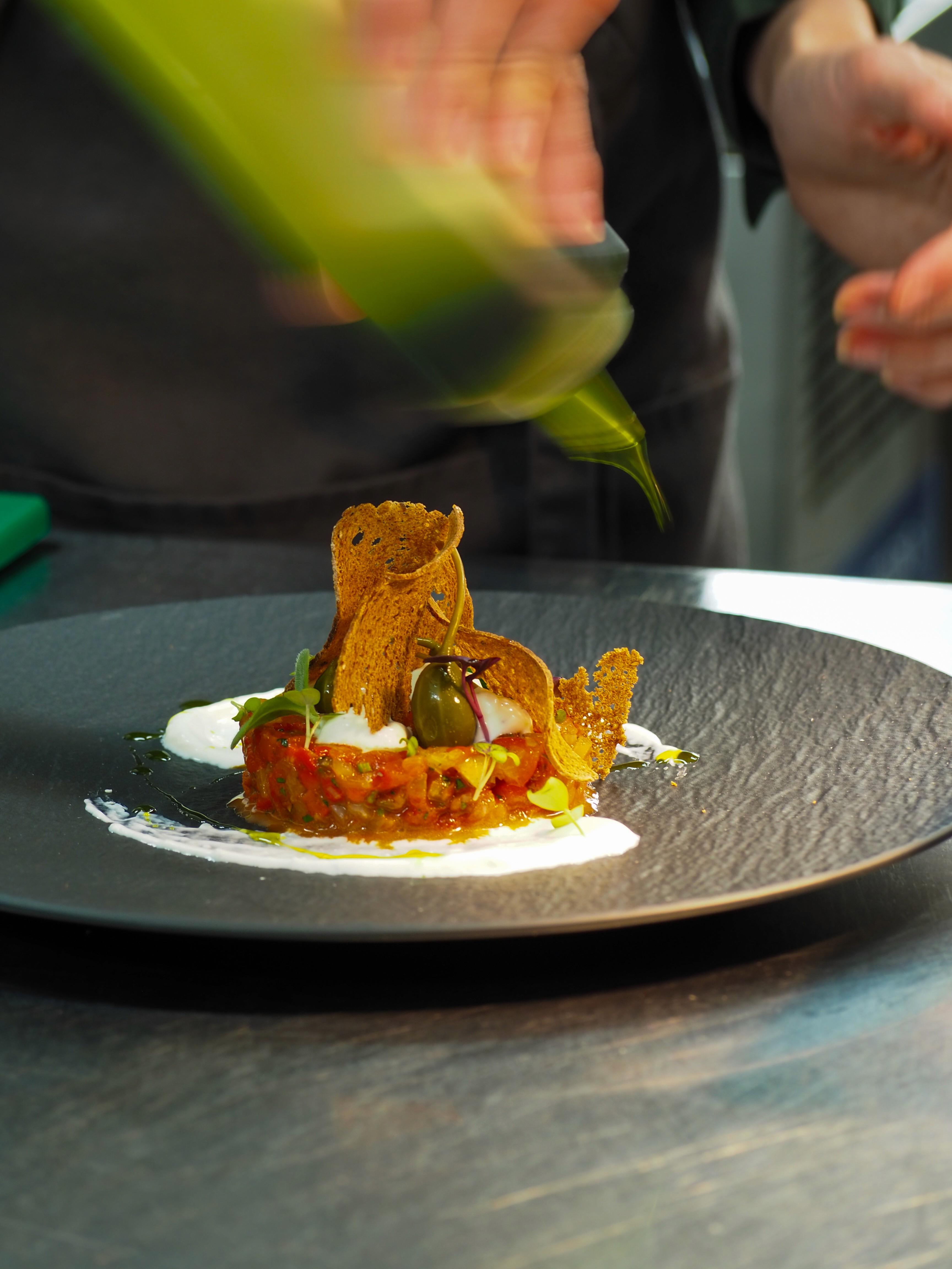 A gourmet dish is being plated on a black plate, with a hand adding a finishing touch from above.