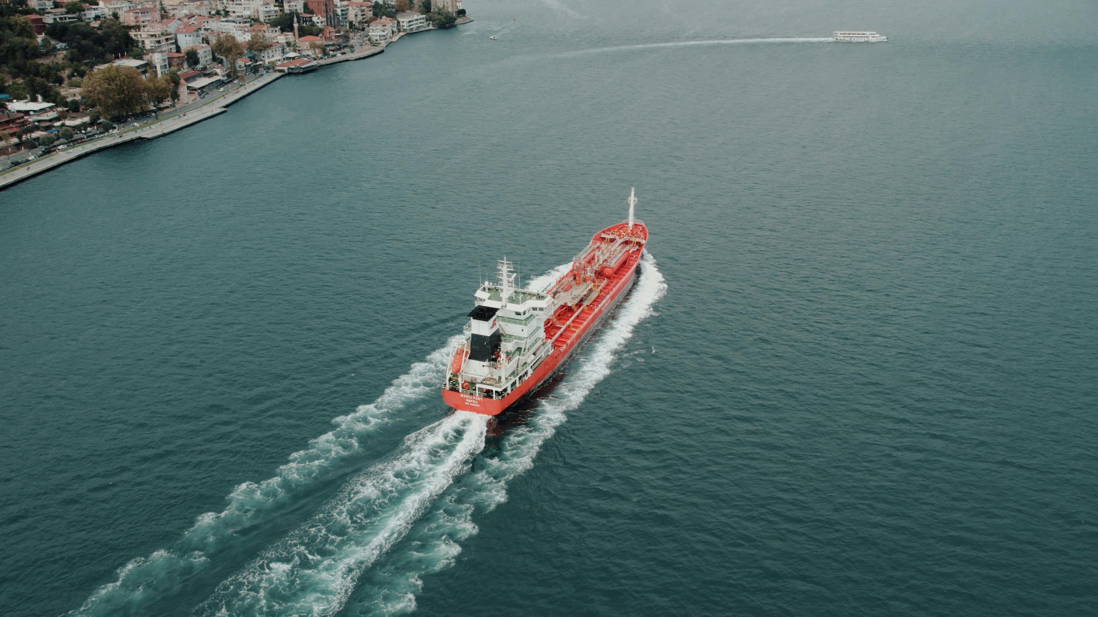 An oil tanker navigates through the tranquil blue waters of the sea, creating gentle waves as it passes a coastal town on the left.