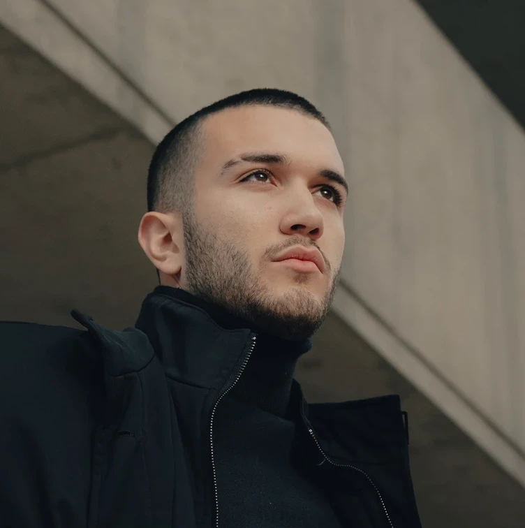 A man with a buzz cut and trimmed beard looks determined, wearing a dark jacket. He stands against a concrete structure, conveying a confident, urban vibe.