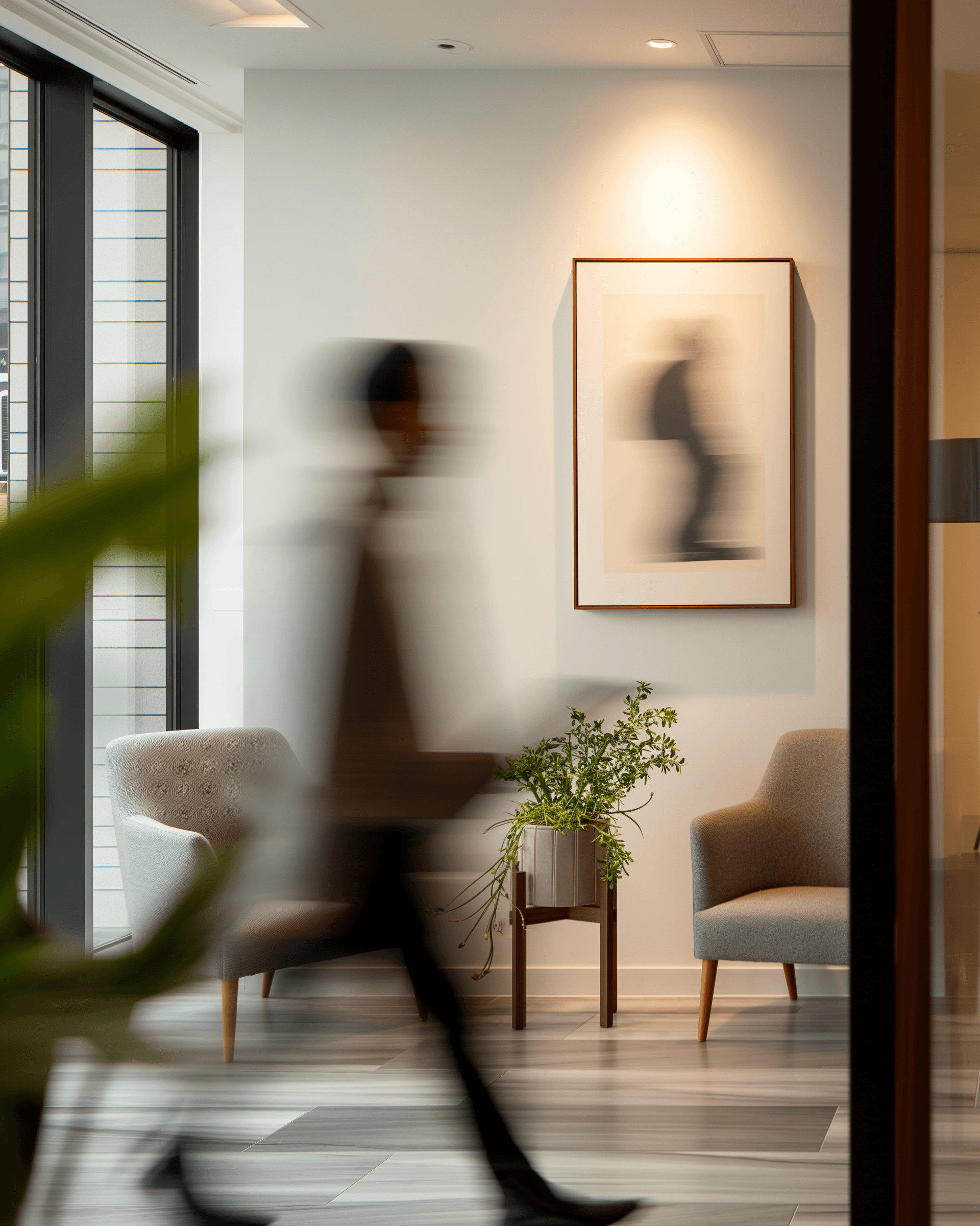 Modern waiting area with gray chairs, a plant stand, abstract art, and a blurred figure walking by, creating a dynamic yet serene vibe.