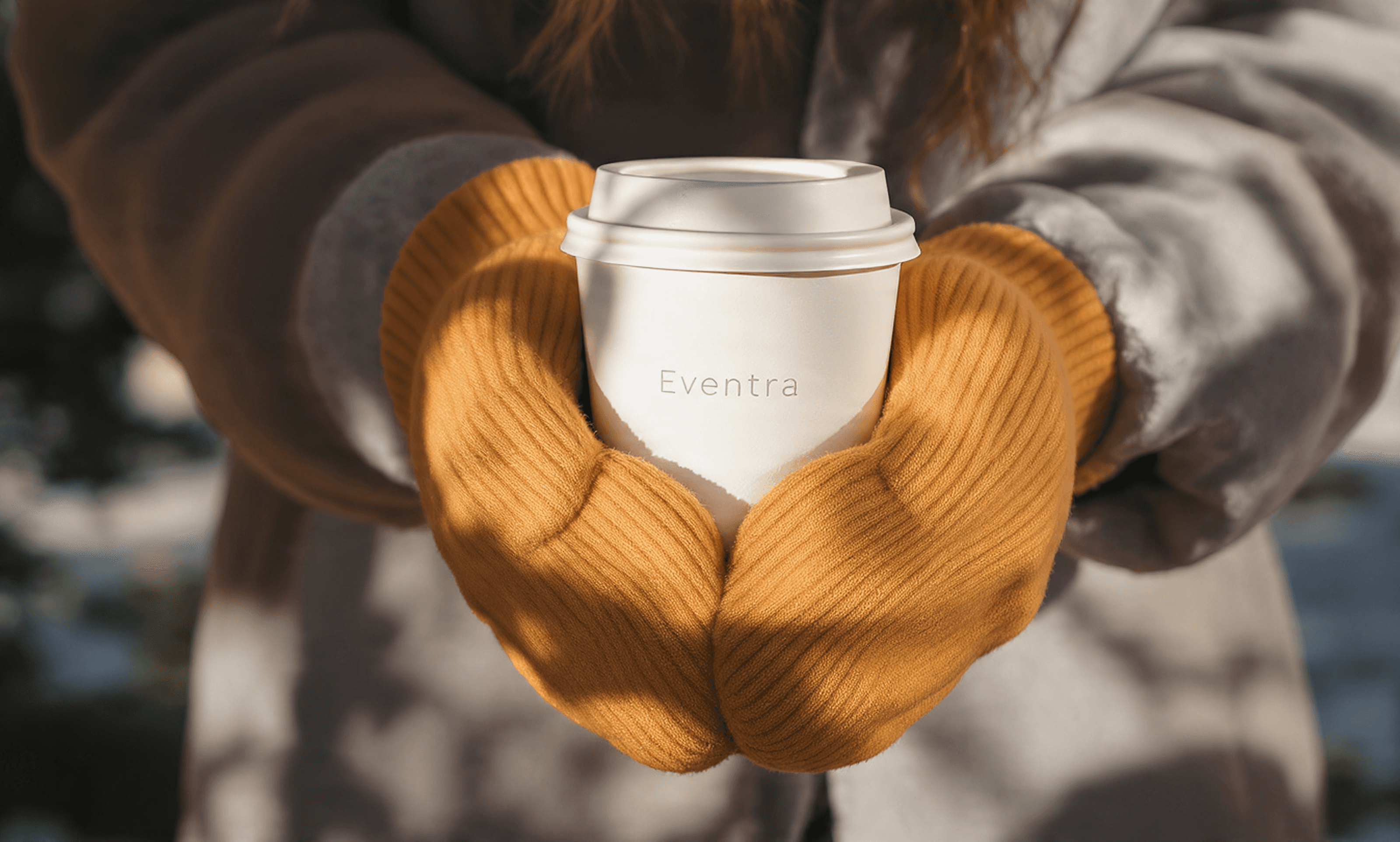 A close-up of hands in yellow mittens holding an 'Eventra' coffee cup.