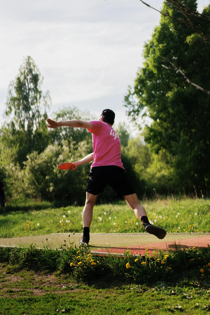 A disc golfer wearing a bright pink Upsi-branded shirt and black shorts is captured mid-throw on a disc golf course. The player is standing on a designated tee pad with one foot lifted, executing a backhand throw with an pink disc. The background features lush green trees and a clear sky, with the sun casting natural light on the scene. The foreground includes grass and yellow wildflowers, enhancing the vibrant outdoor setting.