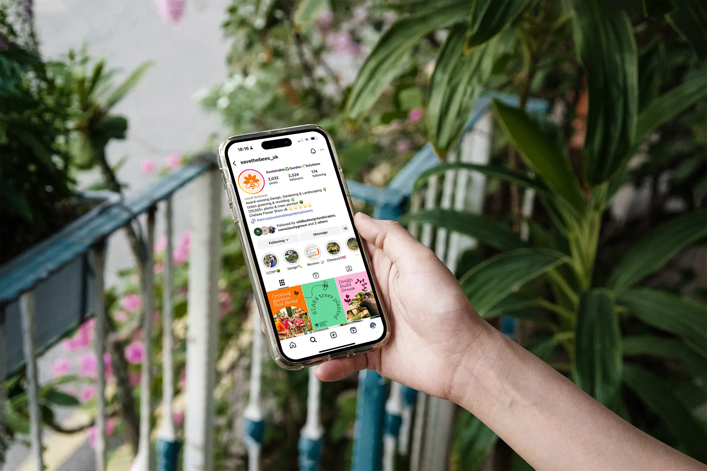 A hand holds a smartphone displaying a colorful app interface, with greenery and stairs in the background.