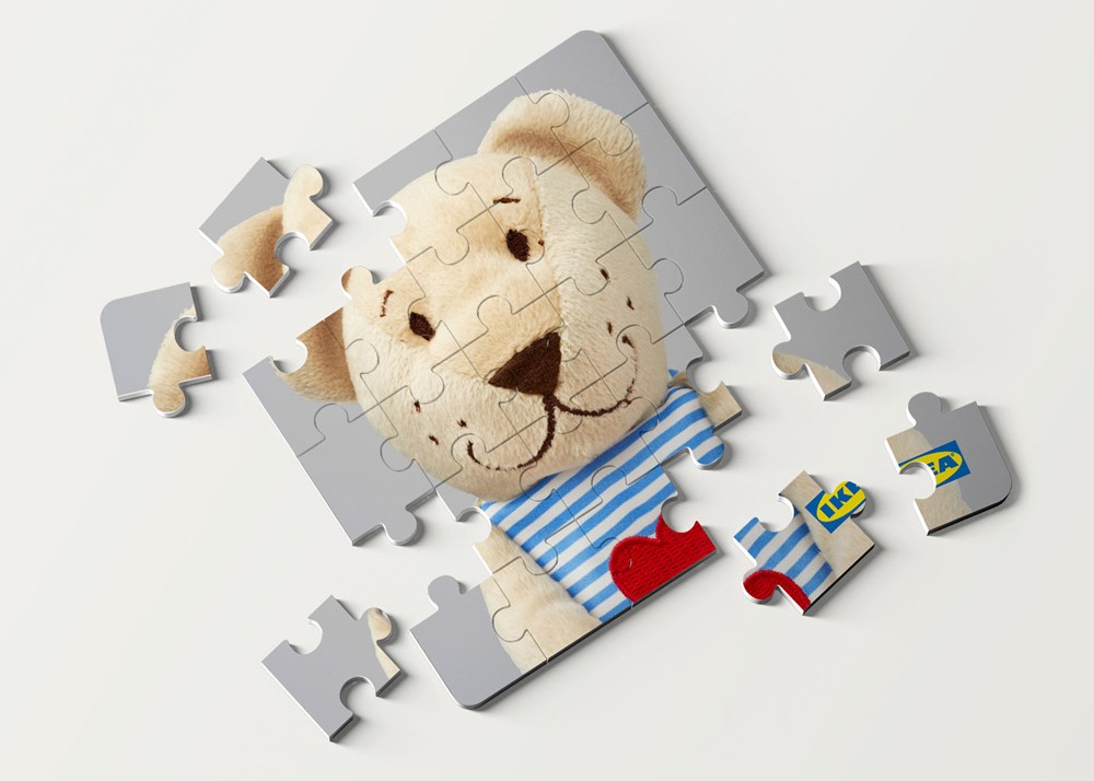 A close-up of a brown bear soft toy surrounded by scattered jigsaw puzzle pieces on a warm surface, from the IKEA Junior Designers children's party activity kit.
