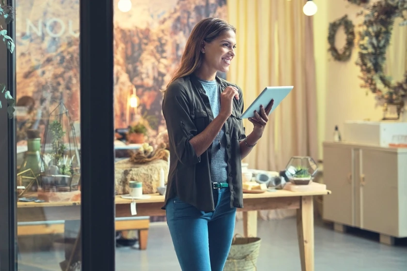A woman is standing inside a store or a boutique, smiling and looking outwards. She is holding a tablet, suggesting she is managing a business or assisting a customer.