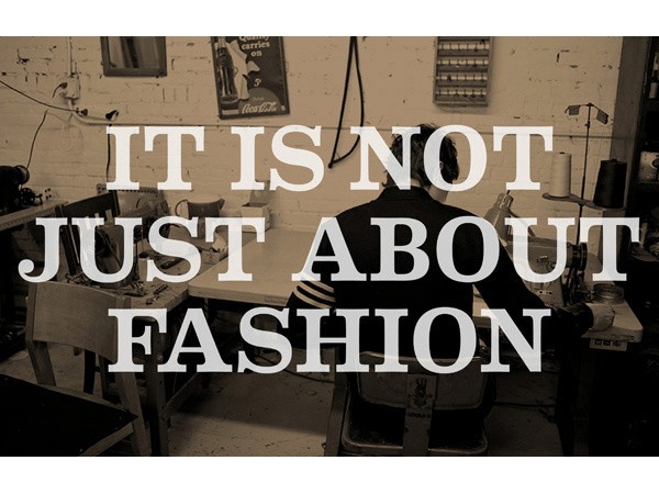 A person sits at a sewing machine in a room with white brick walls. The image is in black and white. Overlaid on the image are the words “IT IS NOT JUST ABOUT FASHION” in large, white, sans-serif font.