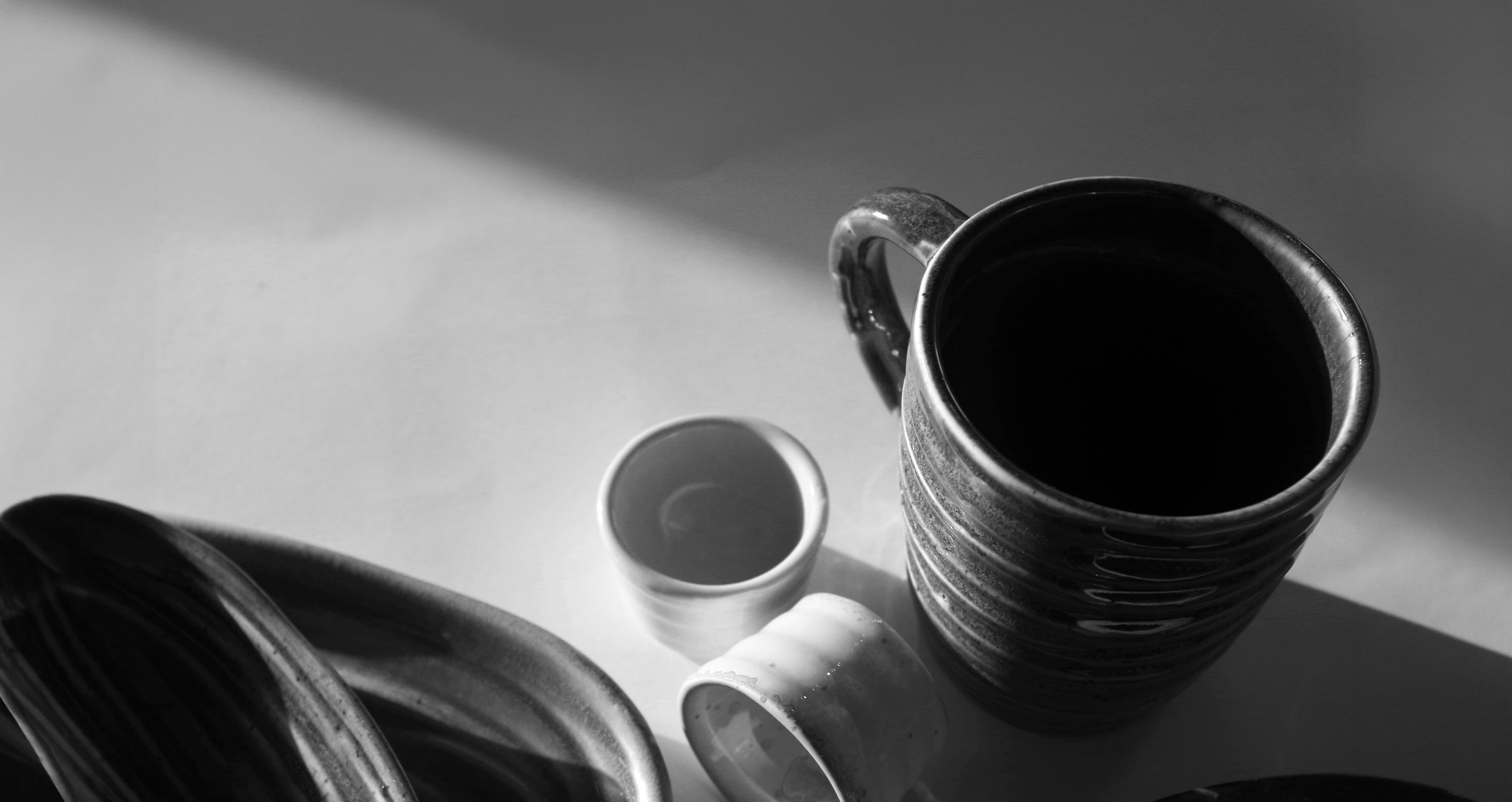 Photographie en noir et blanc d'un grand mug en grès tourné à l'émail noir côtelé, accompagné d'un petit bol et d'une assiette creuse en céramique sombre, éclairés par une lumière naturelle rasante sur surface blanche