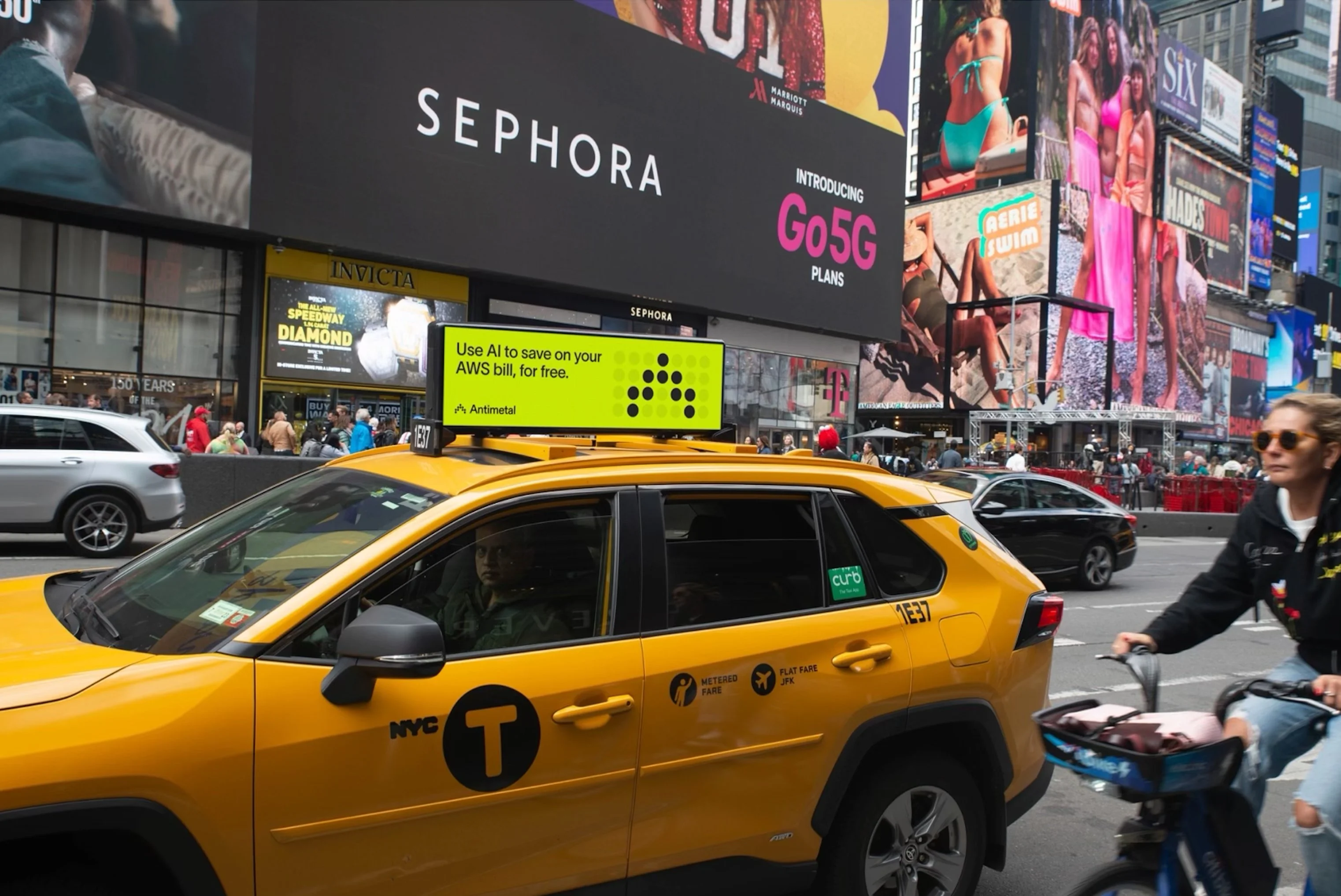 A bustling Times Square scene featuring a yellow NYC taxi with an advertisement on top. The ad reads, 'Use AI to save on your AWS bill, for free. Antimetal.' In the background, there are large billboards including one for Sephora and another for Go5G plans. Pedestrians and a cyclist are also visible in the lively street setting.