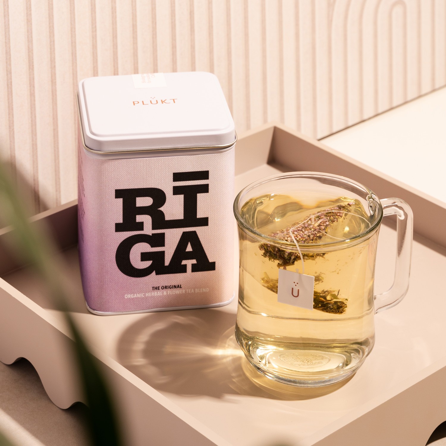 PLŪKT RIGA organiskās tējas iepakojuma fotogrāfija ar stikla krūzīti uz maigas bēšas virsmas — tējas un dzērienu produktu fotografēšana Rīgā.