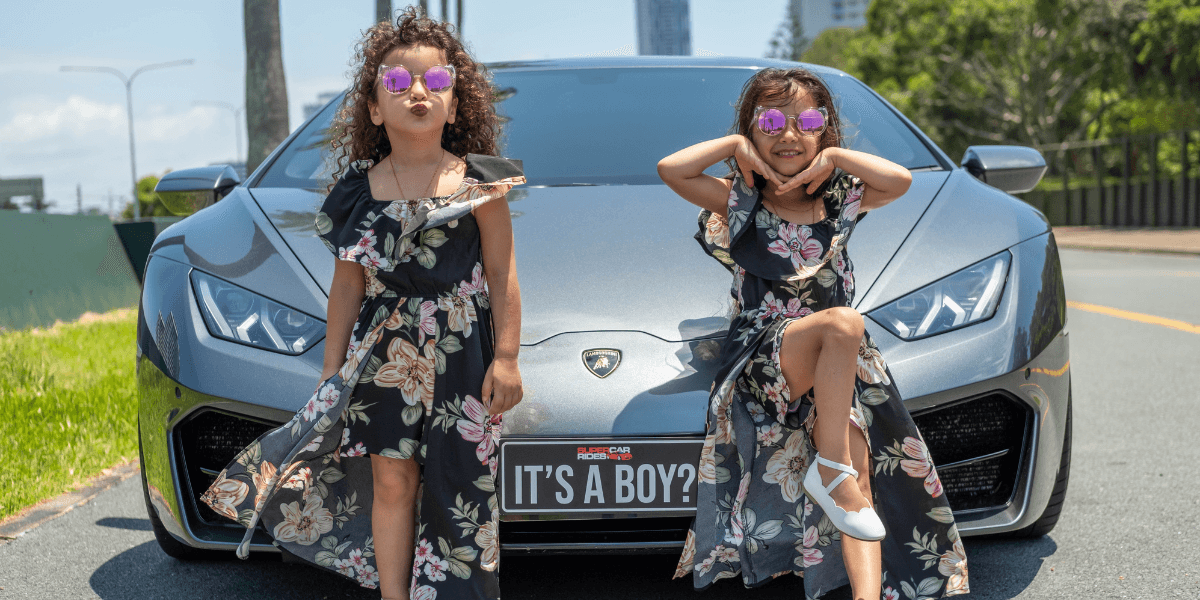 Two young girls in matching floral dresses and sunglasses pose confidently in front of a sleek silver sports car with a "It's a Boy?" license plate.
