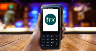A hand holds a Verifone card reader with the TRX logo on the screen.