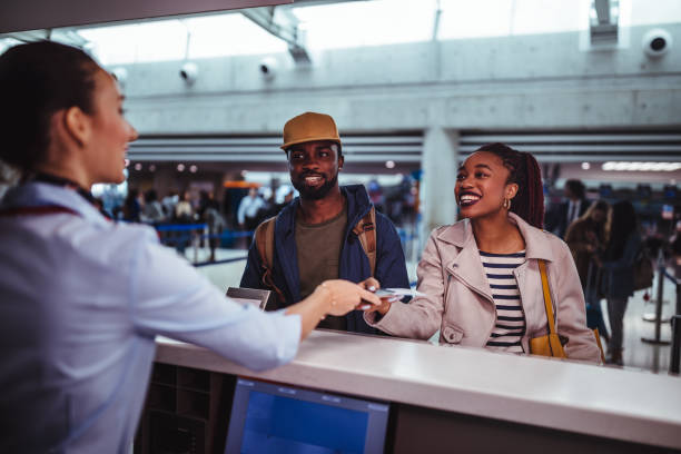 A smiling woman at an airport check-in counter hands her travel documents to a female airline agent. A man in a baseball cap stands next to her, also smiling, as they prepare for their flight.