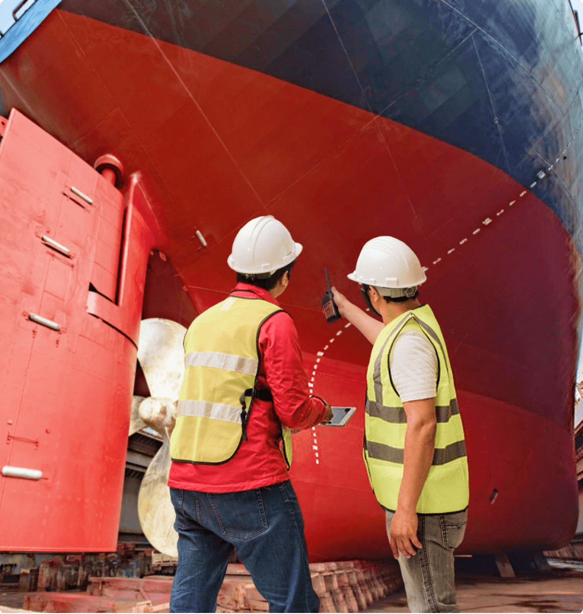 Two workers in hard hats inspect the hull of a large ship in a shipyard, discussing the maintenance details.