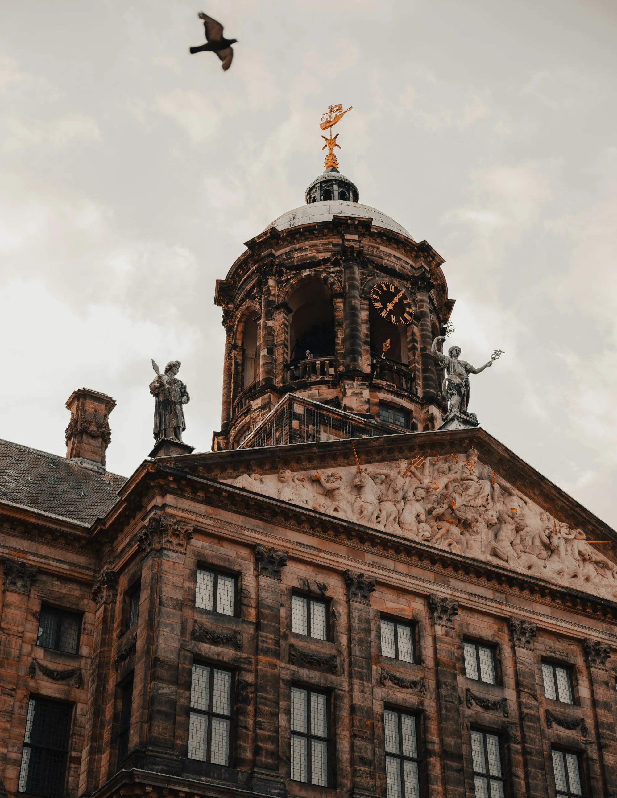 Ornate building with dome and statues, bird flying in cloudy sky, Amsterdam