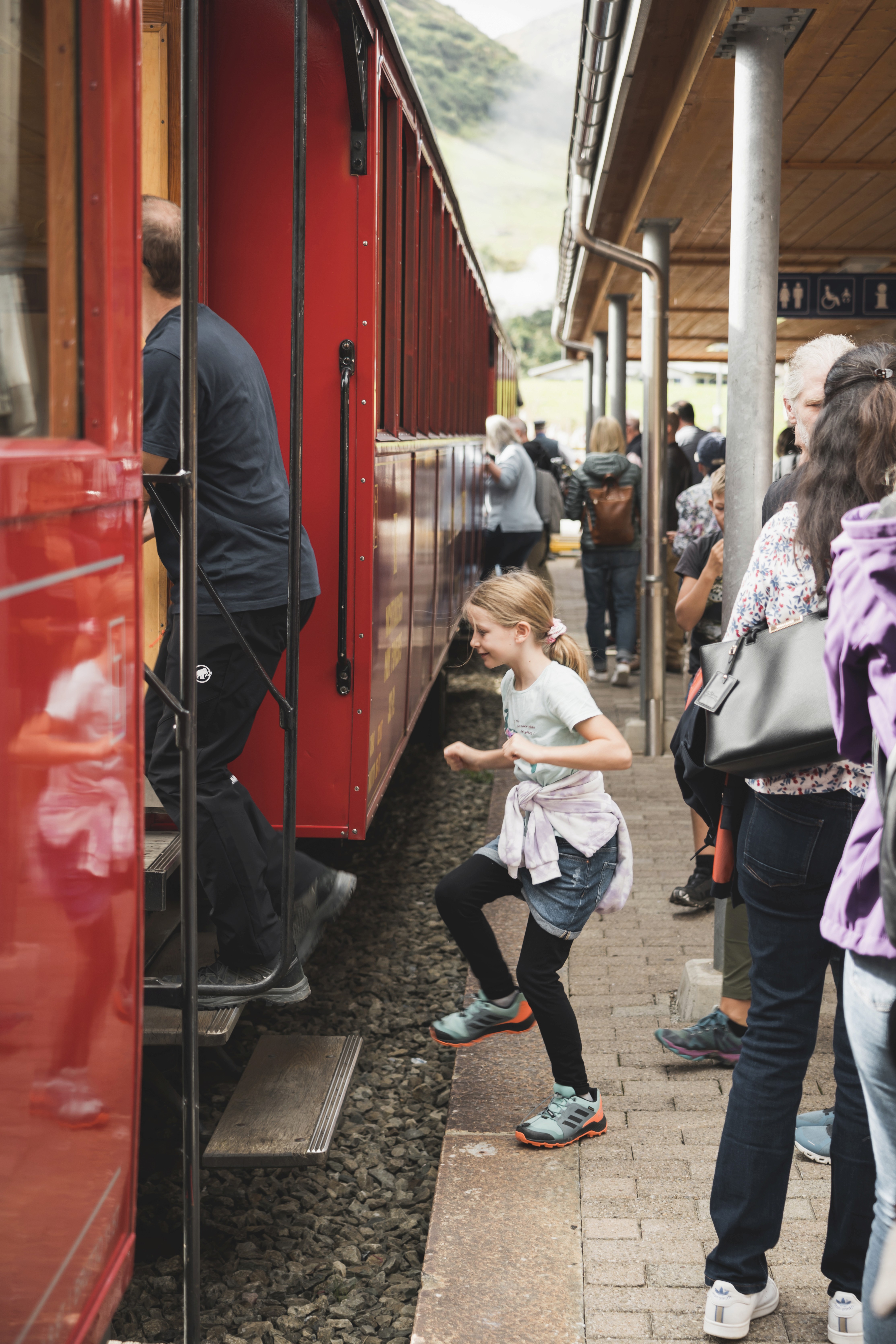 Das Foto fängt eine fröhliche Szene auf einem gepflasterten Bahnsteig ein. Im Zentrum des Bildes hüpft ein Mädchen voller Vorfreude neben einem historischen, leuchtend roten Zugwaggon. Sie trägt ein helles T-Shirt, dunkle Hosen, eine um die Hüfte gebundene helle Jacke und auffällige türkis-orangefarbene Turnschuhe. Links im Vordergrund steigt gerade eine Person in dunkler Kleidung die Stufen in den Waggon hinauf. Im Hintergrund warten weitere Reisende unter der hölzernen Überdachung des Bahnsteigs. Die Szenerie wird im Hintergrund von teilweise nebelverhangenen, grünen Berghängen umrahmt.