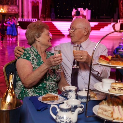 An elderly couple clinks glasses at a table with tea and pastries, while dancers perform in the background on a grand stage.