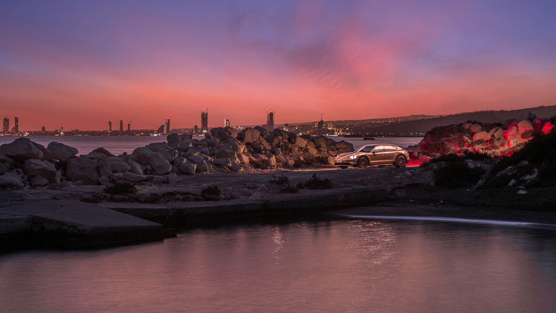 Porsche Taycan Cross Turismo parked among coastal boulders at twilight, seen across calm water, with a city skyline silhouetted under a pink-and-violet dusk sky.