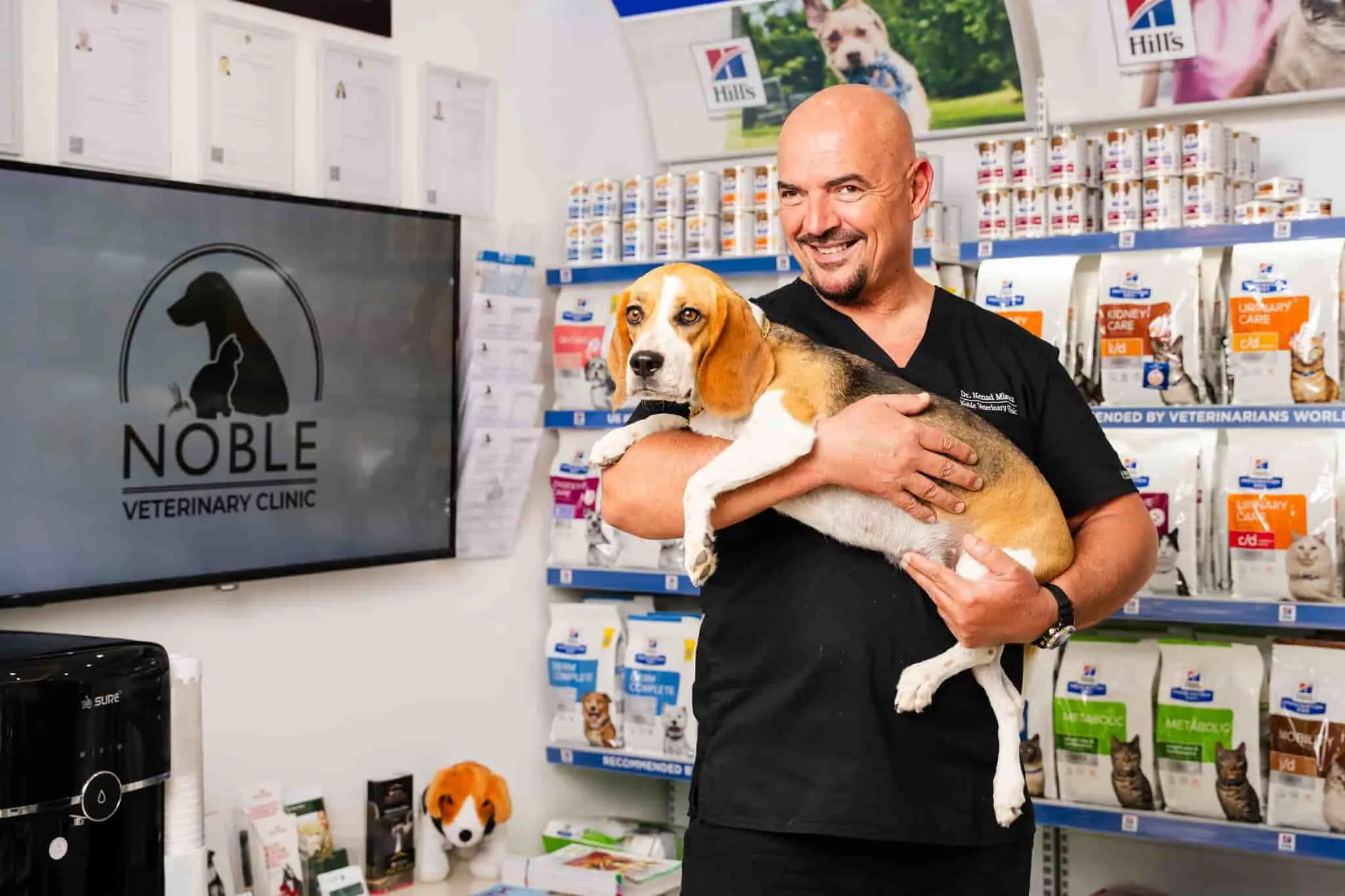 A veterinarian is holding a dog in his arms with a shelf of pet food behind him.