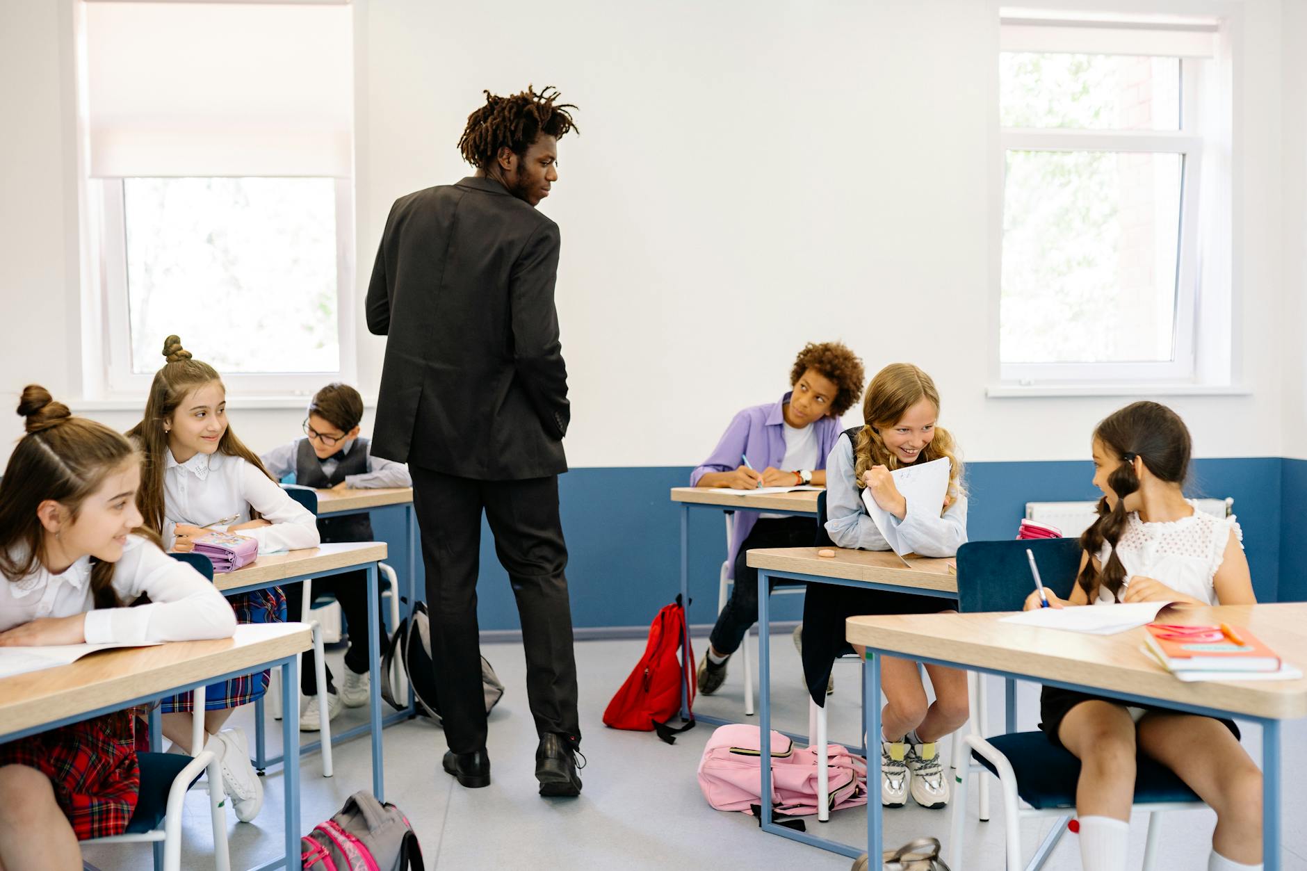 A teacher walking between classroom clusters, observing student groups while holding a digital tablet for assessment.