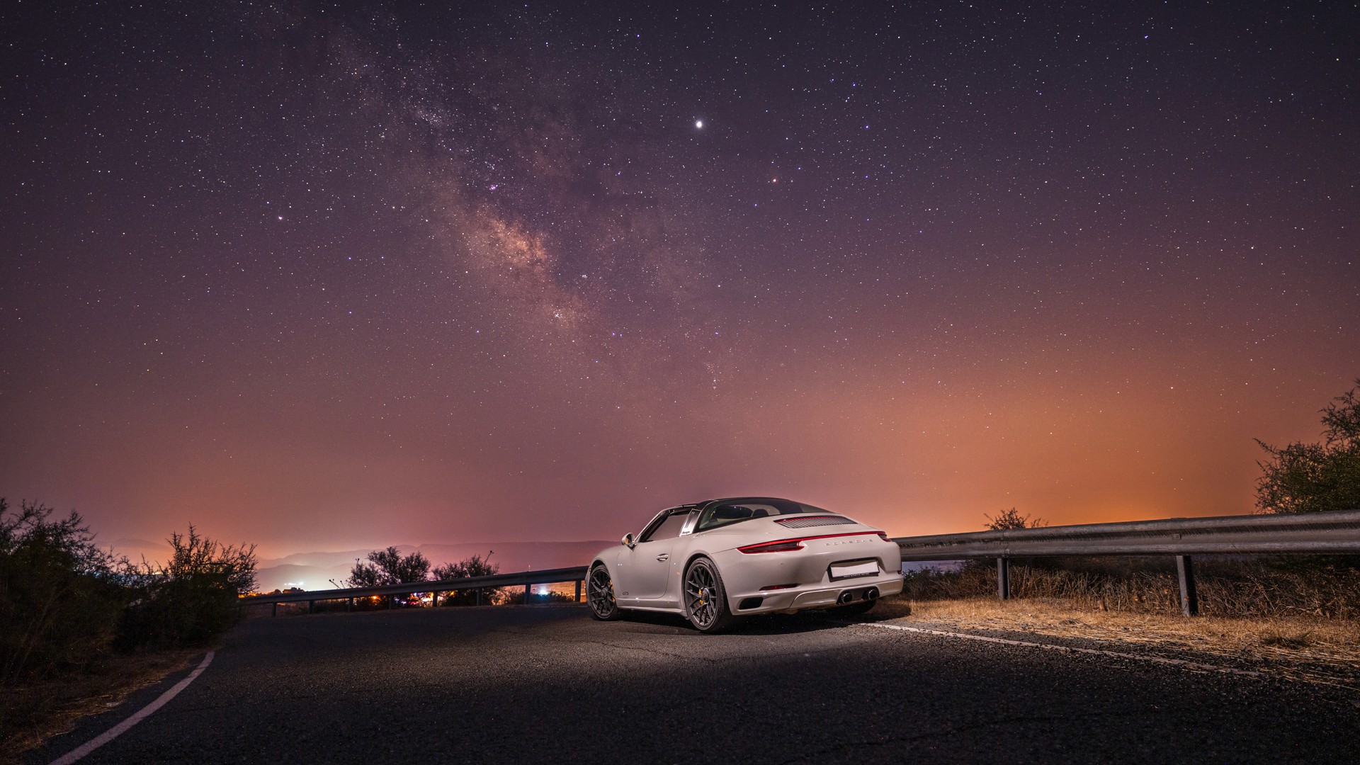 Crayon Porsche 911 Targa GTS parked at a mountain roadside under the Milky Way, rear three-quarter view, the Targa bar and full-width "PORSCHE" light bar visible, with a glowing orange horizon and valley lights below.