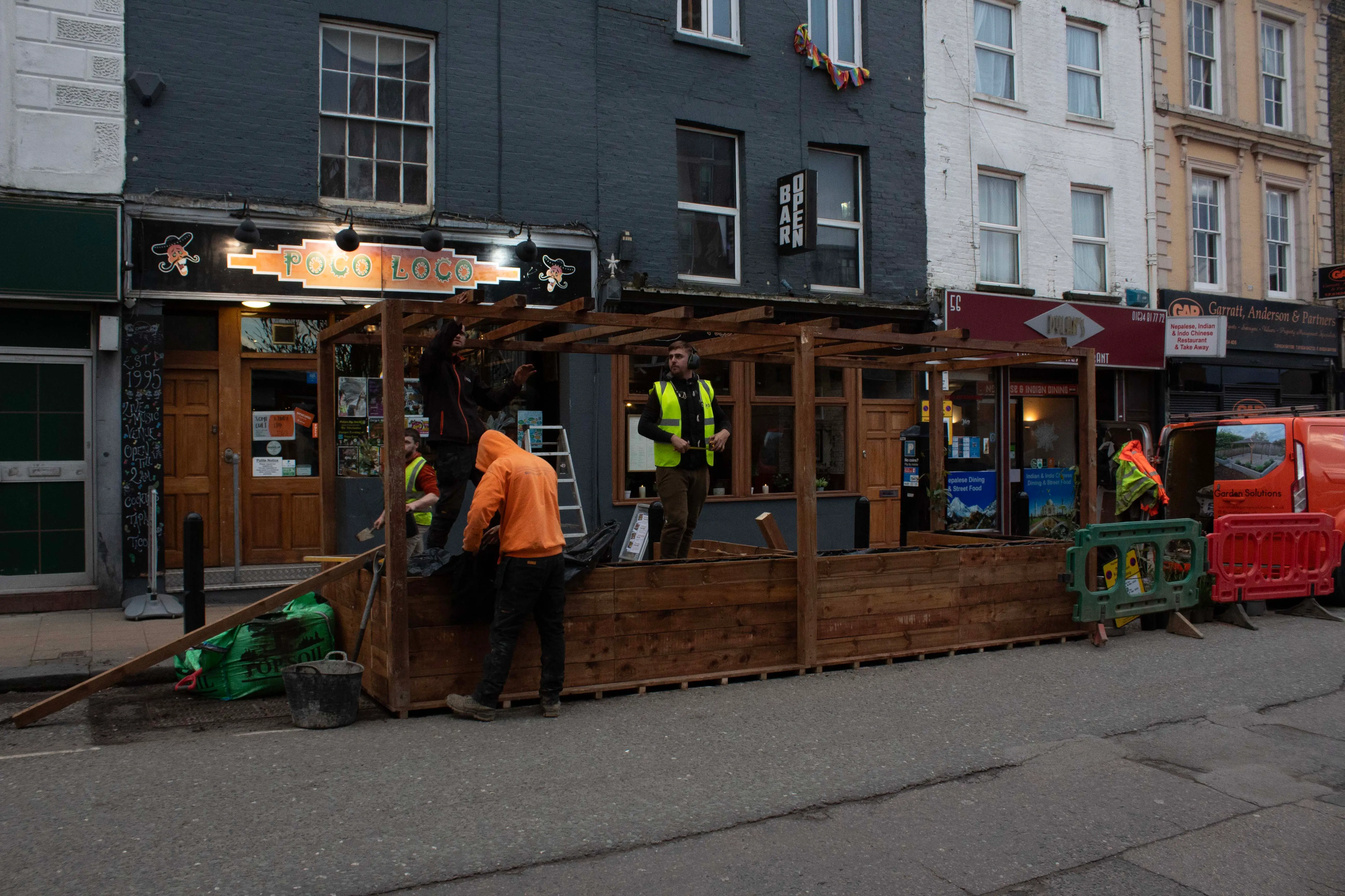 Workers in orange vests are engaged at a wooden construction site on a street, with buildings behind.