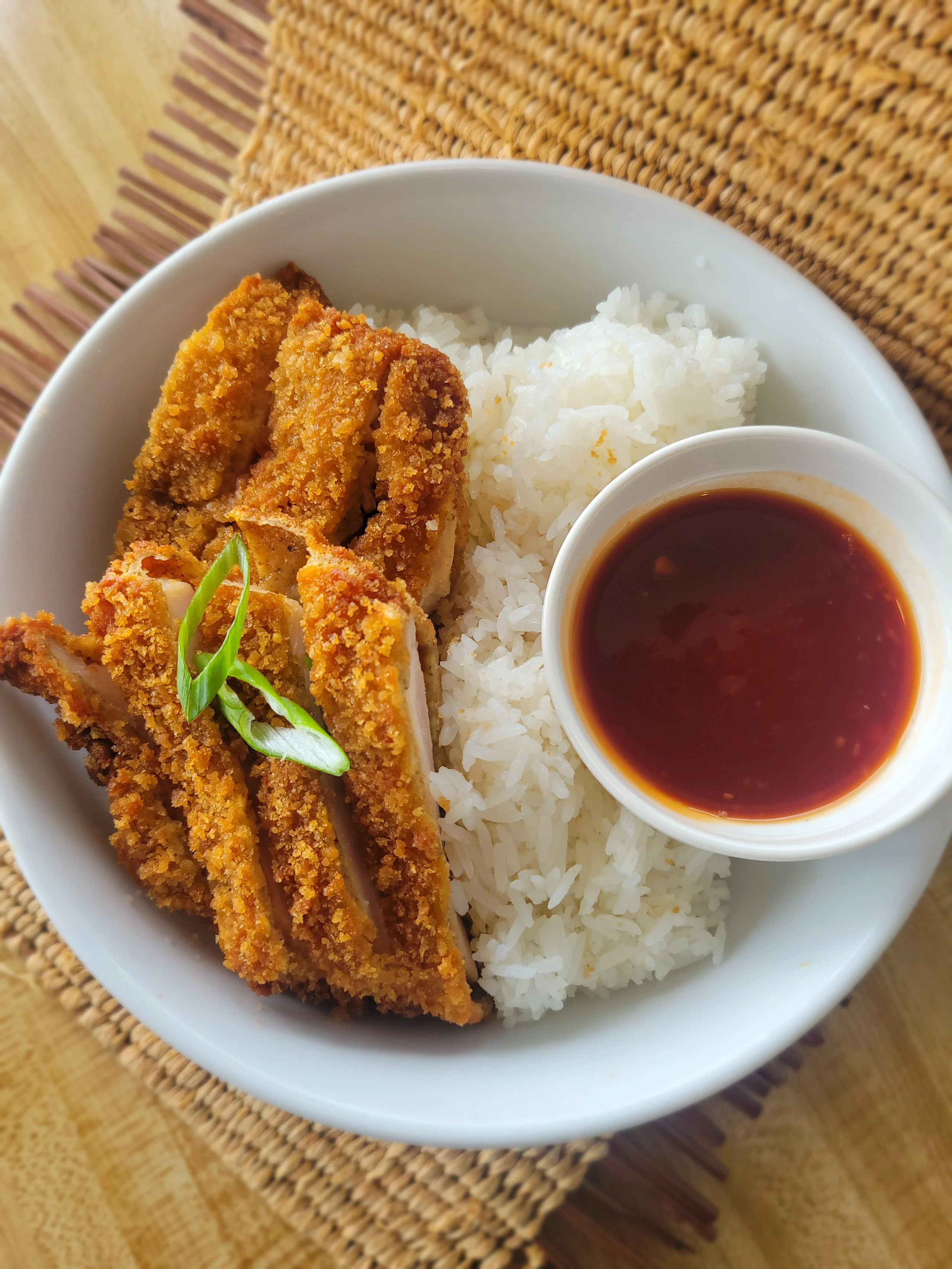 A white bowl featuring sliced, golden-brown crispy chicken katsu served over steamed white rice with a side of dark red katsu dipping sauce.