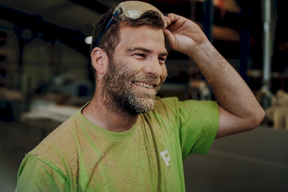 Glücklicher Mann mit Bart und Schutzbrille. Er trägt ein grünes T-Shirt und hat Sägemehl im Gesicht.