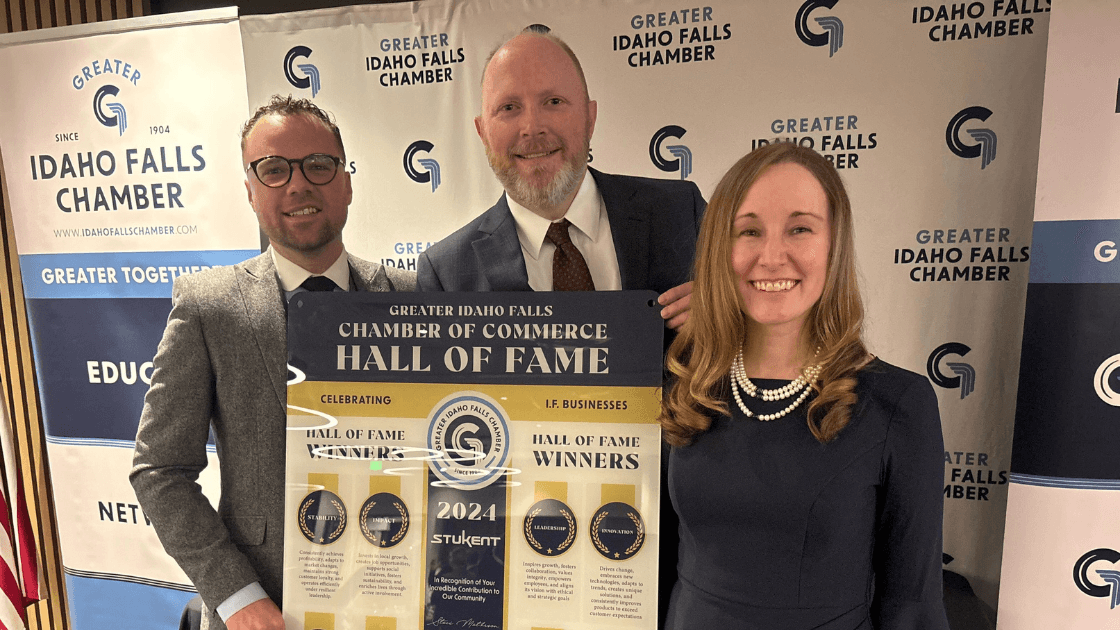 Three people smile while holding a “Greater Idaho Falls Chamber of Commerce Hall of Fame” poster. Behind them is a backdrop with repeating Chamber branding and words like “EDUCATE,” “NETWORK,” and “GROW.”