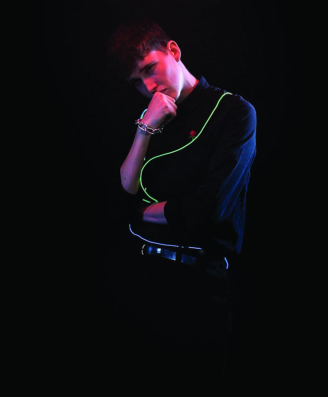A young man with short brown hair stands against a dark background, wearing a black long-sleeved shirt. He holds a thin, glowing green cord around his neck, with his right hand touching his chin and his left arm crossed over his chest. A bracelet is visible on his left wrist, and a microphone is clipped to his belt. The lighting casts red and blue hues on his face and clothing.