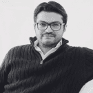 A man with glasses smiles while sitting casually in a sweater against a plain background.